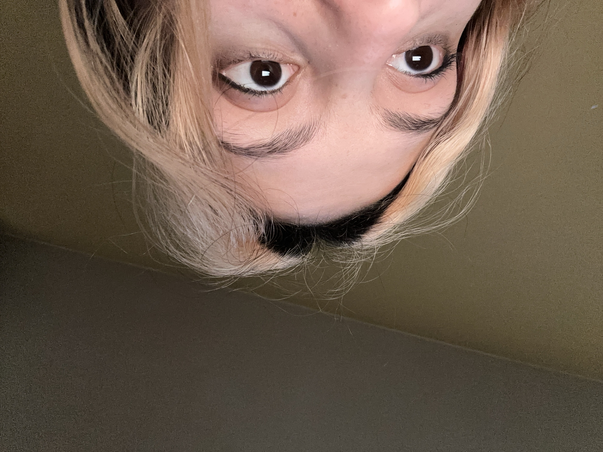 A close-up, downward-facing view of a person's face and hair. The person has fair skin, brown eyes with dark eyeliner, and light brown eyebrows. Their hair is blonde with darker roots. The background is a solid, muted green-brown color. There is no information in the image to determine the location or if the picture was taken in Sofia, Bulgaria.