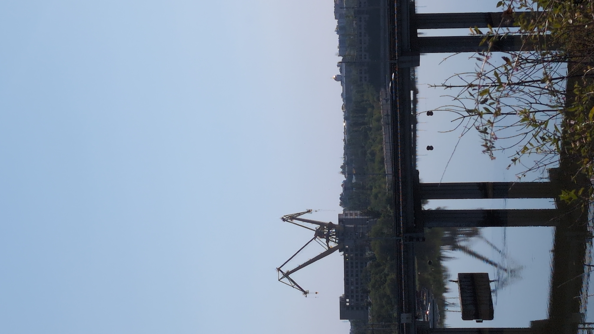 The image depicts a scene in the city of Mykolaiv, Ukraine, likely near a port or industrial area. In the background, a large, yellow gantry crane stands against a clear blue sky, towering over a long, unfinished or abandoned building. To the right of the crane, a cluster of trees lines the horizon, with a hint of urban development beyond, including what appears to be a golden dome on a building.
In the foreground, a modern concrete bridge spans across a body of water. The bridge's pillars are reflected in the calm water, creating a symmetrical image. Small boats are visible in the distance on the water, suggesting it is a river or bay. The foreground also features some lush green foliage, adding a touch of nature to the industrial landscape. The overall impression is one of a functional yet perhaps partially developed or decaying urban environment.