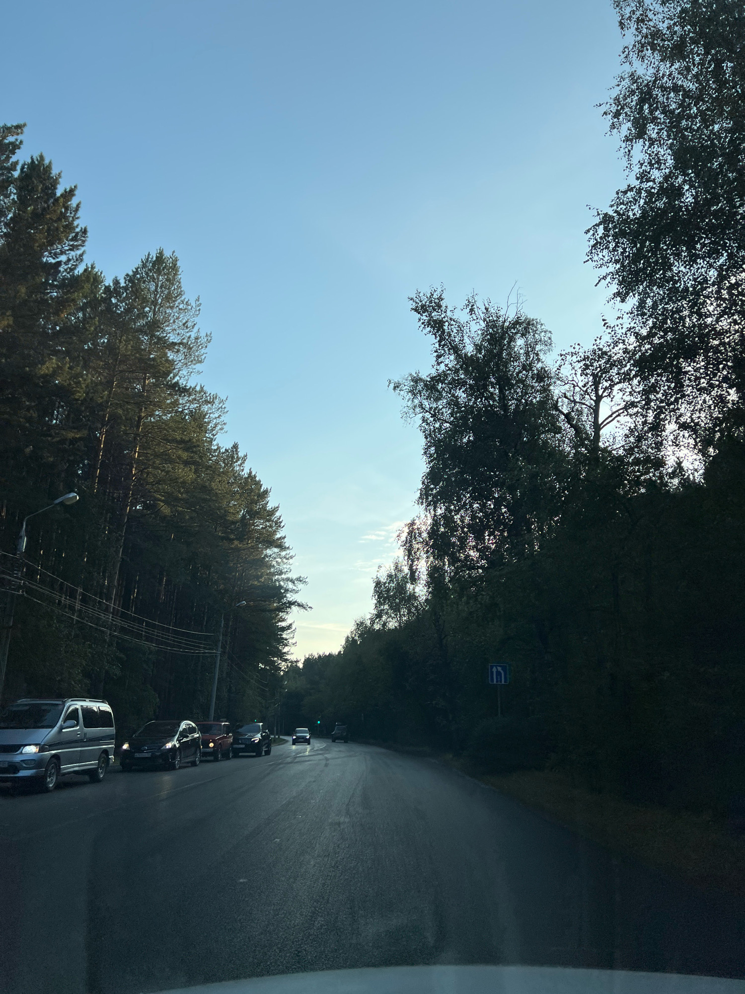 The image shows a view from inside a vehicle driving on a paved road lined with dense evergreen and deciduous trees on both sides. The sky is a clear, pale blue with a few wispy clouds. Several cars are driving in the same direction as the viewer, with one van and a line of cars visible ahead. There are also utility poles and wires on the left side of the road. A road sign indicating a merge or lane change is visible on the right side of the road. The scene appears to be on the outskirts of a city, likely during daylight hours. Based on the trees, road type, and general atmosphere, this could be a location in or around Krasnoyarsk, Russia, possibly on a route leading into or out of the city through a forested area.