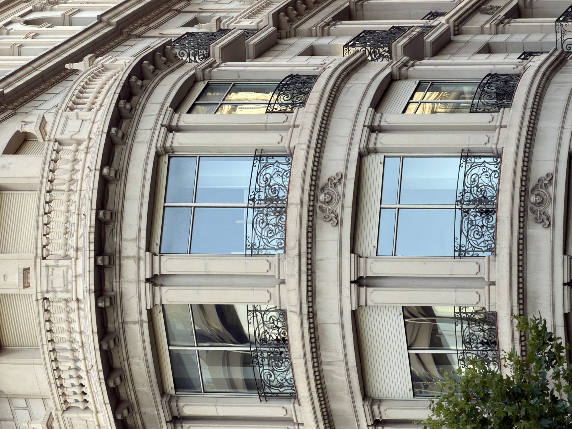 The image shows a detail of a curved stone building facade in Barcelona, Spain. The building has multiple stories with large windows and ornate balconies. The windows are mostly glass, reflecting the sky and surrounding architecture, while some have roll-down shutters. The balconies feature intricate wrought-iron railings and are supported by stone brackets. Decorative stone carvings, including what appear to be faces, adorn the facade between floors. The overall style suggests a building from the early 20th century, likely in a classical or modernist architectural style common in Barcelona.