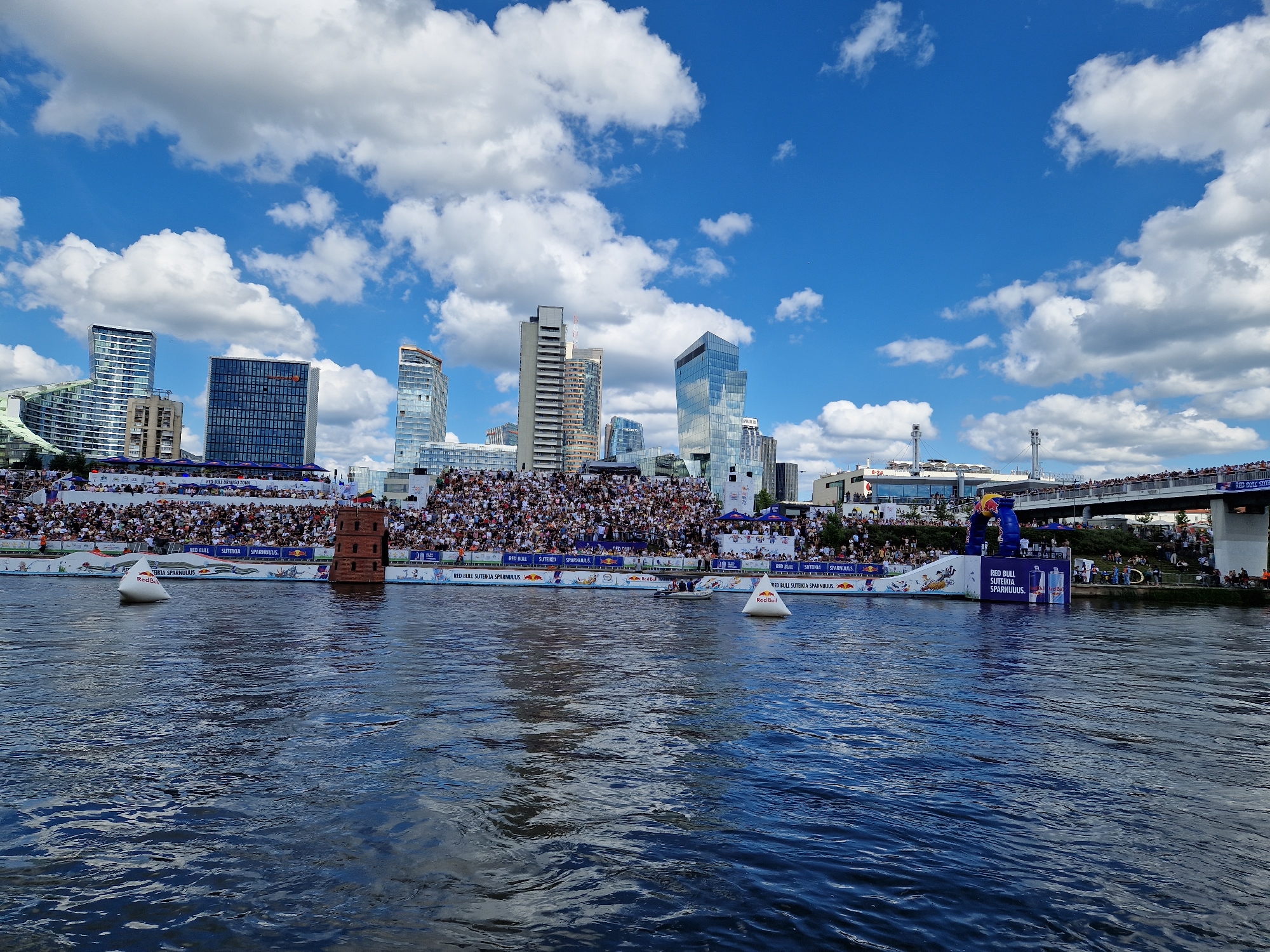 The image captures a wide view of an outdoor event taking place on the Neris River in Vilnius, Lithuania, likely a Red Bull sponsored competition given the prominent branding. The scene is divided between the dynamic water in the foreground and a bustling event area in the background.
In the foreground, the river's surface is a deep blue, rippling with sunlight, suggesting a bright, clear day. Several white buoys, one distinctly branded with "RED BULL," are visible in the water, marking the course of the event. A small motorboat is also present.
The background reveals a vibrant city skyline featuring modern skyscrapers with glass facades, interspersed with older architectural styles. A large crowd of spectators is densely packed on tiered seating that runs along the riverbank, facing the water. Above the seating, the city's buildings provide a dramatic backdrop.
Dominating the middle ground of the event area is a distinctive, red, tower-like structure, possibly a competition platform or landmark. Red Bull branding is pervasive on banners and structures along the riverbank, including what appears to be a ramp or diving platform on the right side of the frame, adorned with Red Bull logos and inflatable figures. The overall atmosphere suggests a well-attended, energetic public event.