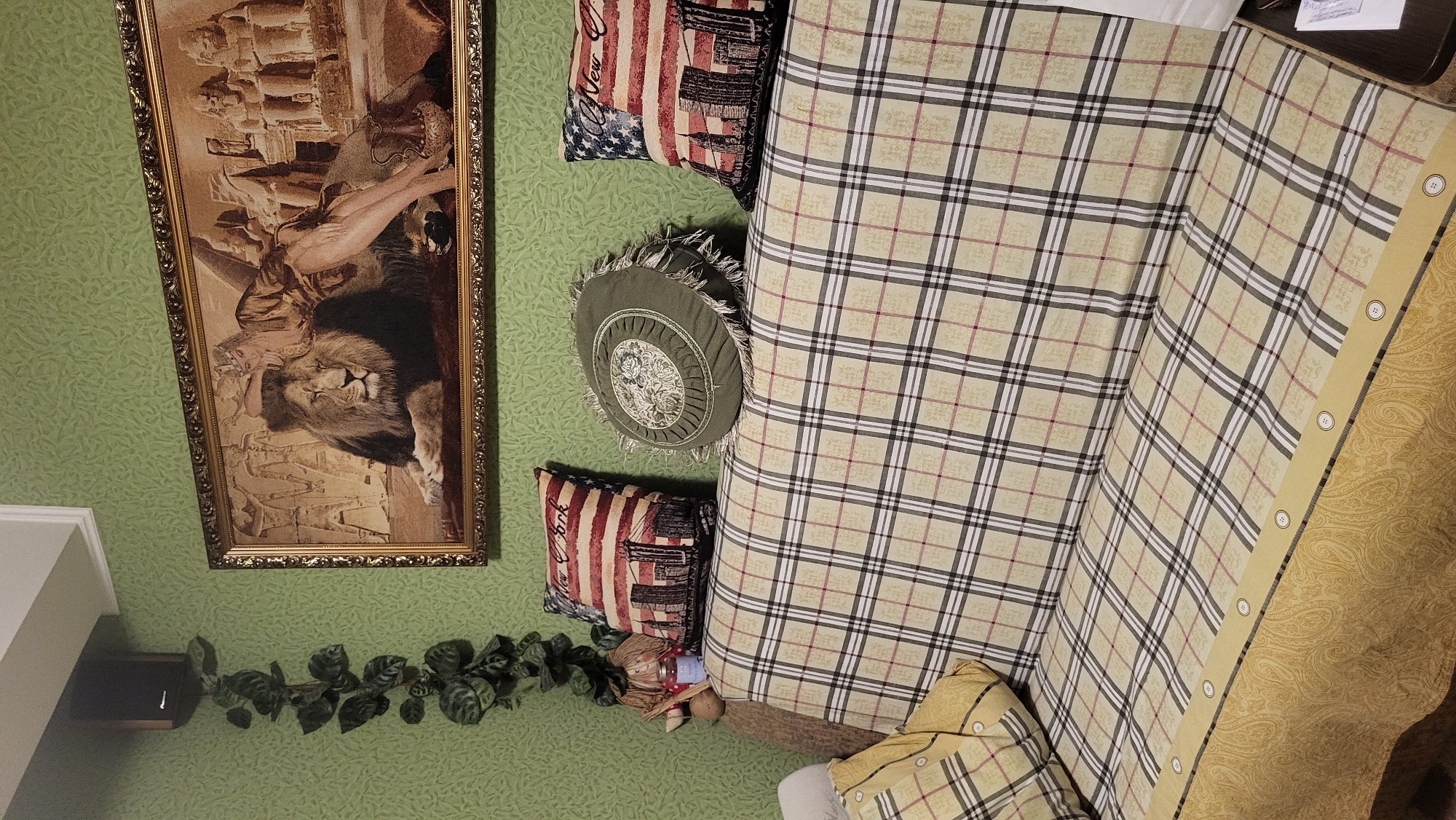 The image depicts an indoor scene, likely a living room, with a prominently featured sofa covered in a beige and black plaid fabric. The sofa has several decorative pillows: two rectangular pillows with an American flag and New York City skyline design, and one round, green pillow with a floral pattern and fringe. Behind the sofa, a tapestry hangs on a light green, textured wall, depicting a lion with a woman and Egyptian-style statues. To the left of the tapestry, a dark speaker is mounted on the wall, with a trailing artificial plant hanging beside it. The overall impression is one of a decorated living space. The location in Smila, Ukraine is not visually ascertainable from the image.