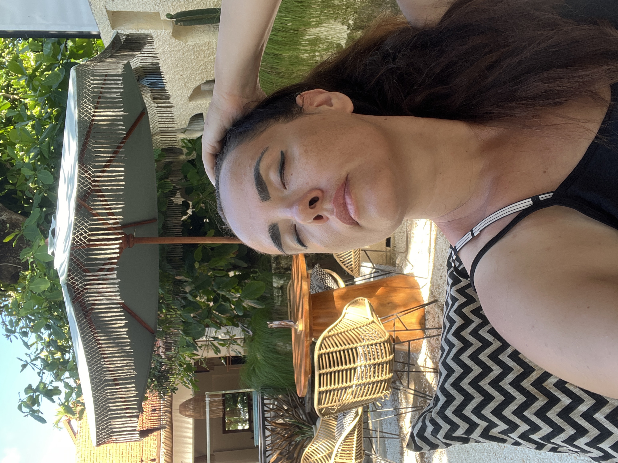 A medium shot captures a woman posing for a selfie outdoors, her eyes closed and hand resting on her head. She is seated on a textured cushion with a black and white chevron pattern, possibly on a chair or sofa. In the background, a circular wooden table with wicker chairs surrounds it, suggesting an outdoor dining or lounge area. A decorative umbrella with fringes is positioned above the table, and lush green foliage is visible, indicating a tropical setting. The architecture suggests a stylish resort or cafe. The woman has long, dark, wavy hair and is wearing a black tank top with thin straps. The lighting indicates it is daytime, with sunlight illuminating the scene. The overall impression is one of relaxation and enjoyment in a resort-like environment.