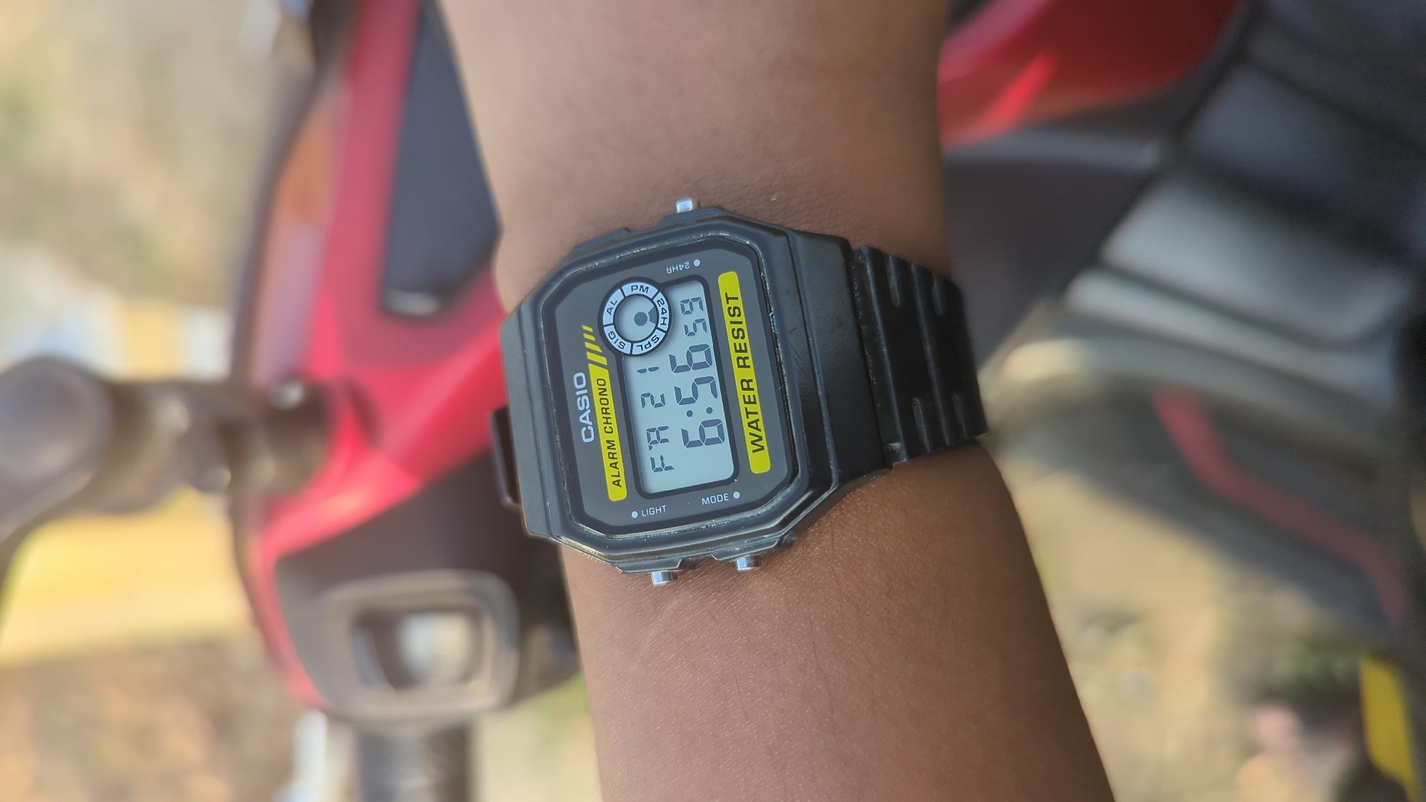 A close-up, eye-level, outdoor shot shows a person's left wrist wearing a black Casio digital watch. The watch face displays the time 6:56:59, the date FR 21, and indicates "ALARM CHRONO" and "WATER RESIST." The background is blurred, suggesting a focus on the watch. The scene is likely in Semarang, Indonesia, given the presence of a red and black scooter, which are common in the region. The lighting suggests it is daytime.