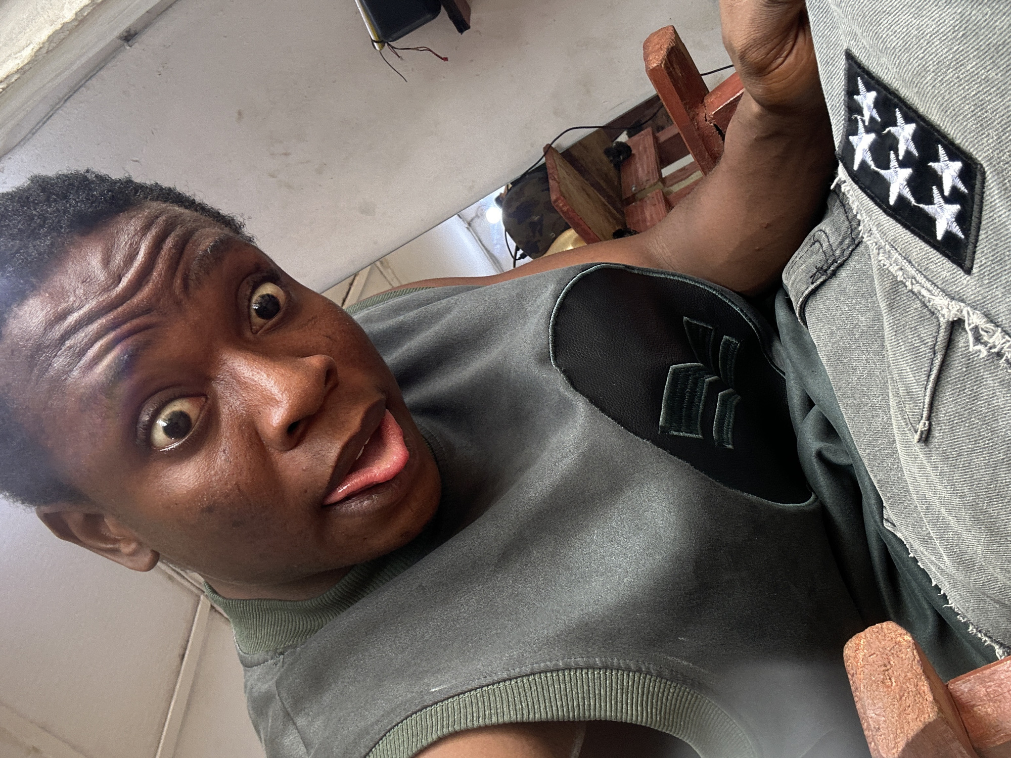 A person is shown from a low angle, close-up, with their tongue sticking out and eyes wide, indicating a playful or silly expression. The person is wearing a gray shirt with a collar and a darker gray or black piece with three striped decorations on the shoulder area. A light gray denim jacket or shirt is also visible, featuring a patch with a black background and six white stars. The background is blurry but appears to be an indoor setting with a white wall and some dark objects. A wooden chair is also visible in the background. The location is Unknown, Unknown.