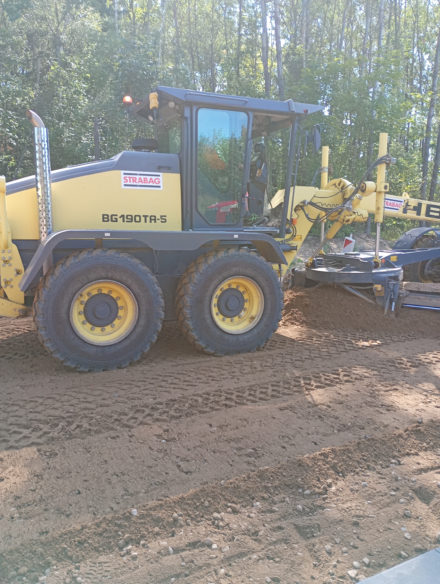 The image shows a yellow road grader with the model number "BG190TA-5" and the company logo "STRABAG" on its side, operating on a dirt surface. The grader is positioned in what appears to be a construction or landscaping site. The background consists of a dense line of deciduous trees under a bright, sunny sky. The ground beneath the grader shows tire tracks and loose soil, indicating recent activity. The grader is in the process of leveling or moving the soil with its blade. The location is specified as Grabówka, Poland.