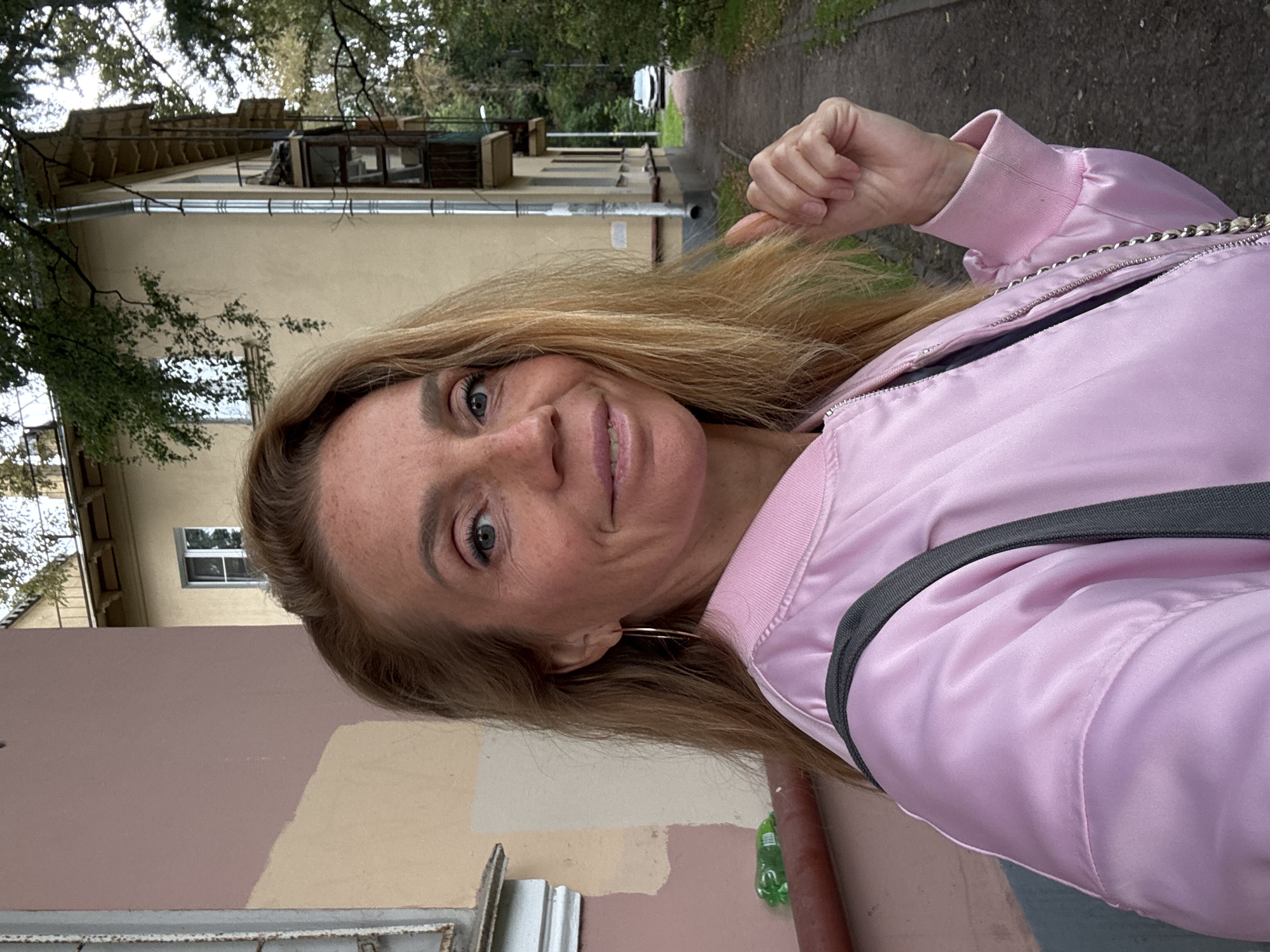 A woman with blonde hair, freckles, and blue eyes takes a selfie outdoors. She is wearing a pink bomber jacket and gold hoop earrings. Behind her, a building with yellow and pink paint is visible, along with trees and a paved walkway. A white car is parked on the street in the background. The scene appears to be in a residential area of Saint Petersburg, Russia, possibly during autumn due to the fallen leaves on the ground.