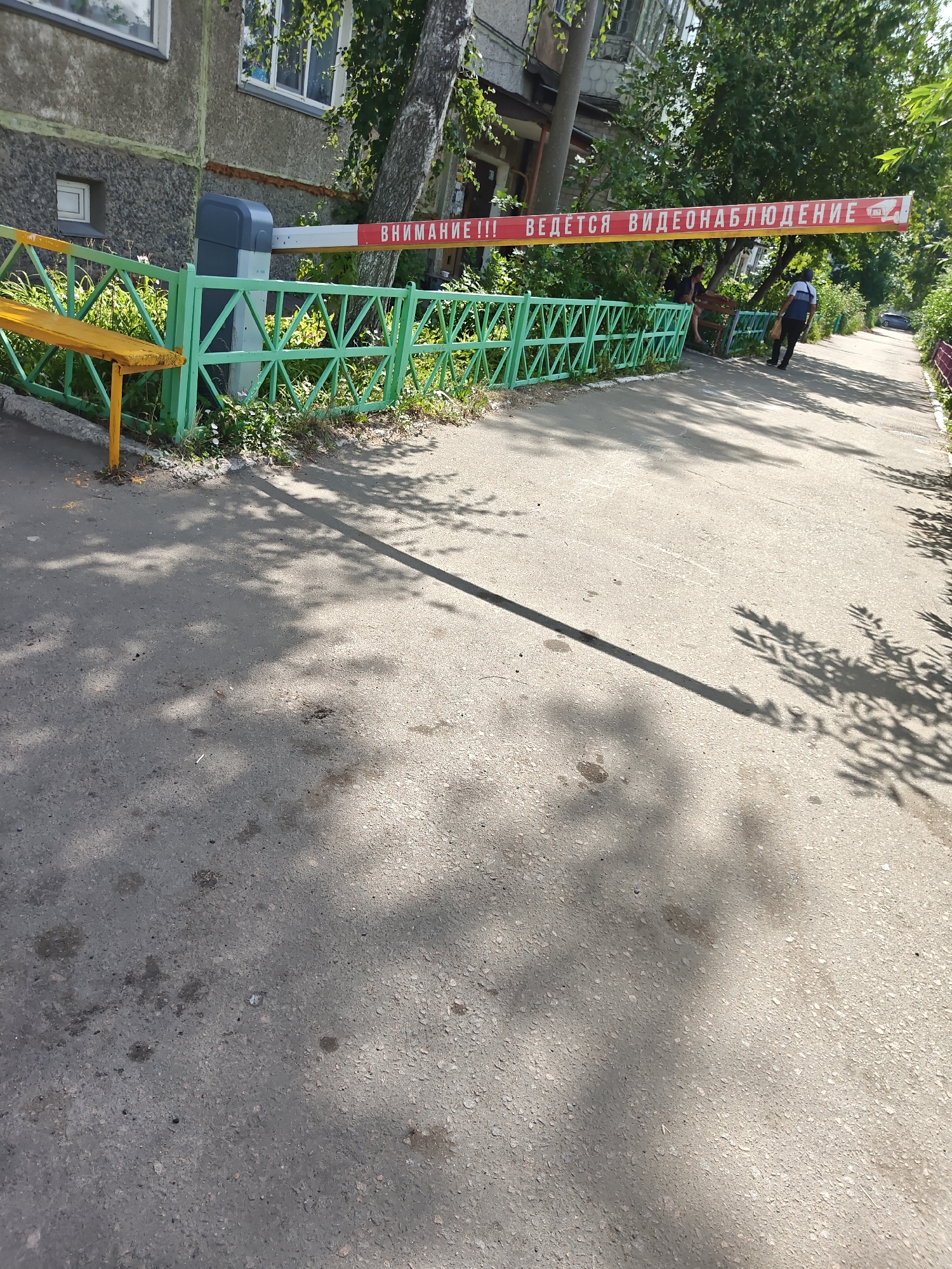 The image depicts an outdoor scene with a red and white striped barrier across a paved pathway. On the barrier, text in Russian reads "ВНИМАНИЕ!!! ВЕДЕТСЯ ВИДЕОНАБЛЮДЕНИЕ", which translates to "ATTENTION!!! VIDEO SURVEILLANCE IS BEING CONDUCTED". A green metal fence with a yellow bench is positioned to the left of the barrier. A man in a white shirt and dark pants, carrying a dark bag, walks away from the viewer towards the right side of the image. Another person, partially visible and sitting, is also on the other side of the fence. The pathway is flanked by trees and foliage, casting shadows on the ground. The background shows a building and a tree-lined area. The scene appears to be in a residential or semi-public area. The location in Petropavl, Kazakhstan is not discernible from the visual information in the image itself.