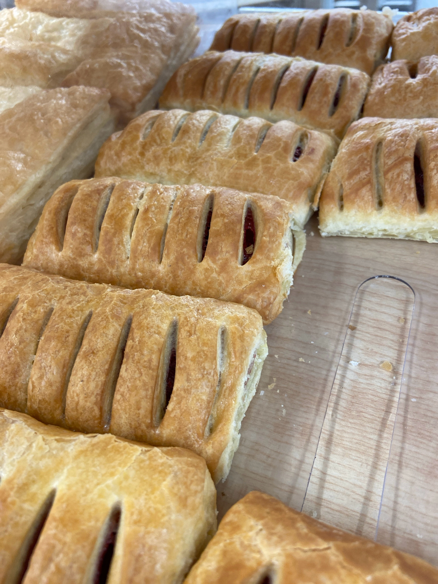 The image shows a close-up view of several golden-brown pastries arranged on a light-colored wooden surface. The pastries appear to be filled, with visible red fruit filling peeking through the slashes on their tops. Some pastries are rectangular, while others are more triangular. In the background, more pastries are stacked, and a clear plastic utensil or edge is visible on the right side of the frame. The lighting suggests an indoor setting, possibly a bakery or display case. There are no people or animals present in the image. The image does not contain any information that would allow for the determination of its location in Balashikha, Russia.