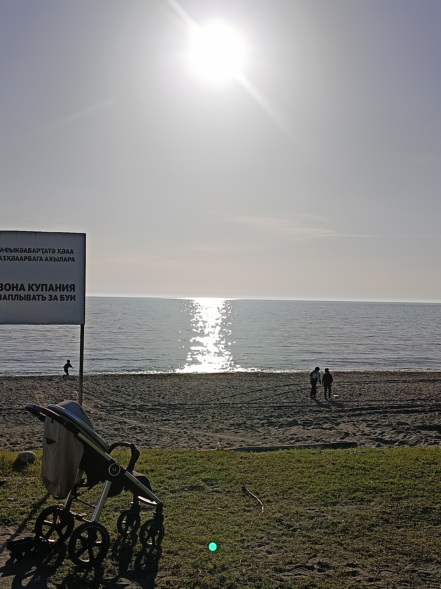The image shows a beach in Gulrypsh, Abkhazia on a sunny day. In the foreground is a baby stroller parked on a grassy area bordering the beach. Behind the stroller, there is a sandy beach with a sign on the left that appears to be in Russian and another language. In the distance, there are two people walking on the beach, and another person is near the water. The sea extends to the horizon where it meets the sky. The sun is high in the sky, causing a reflection on the water.