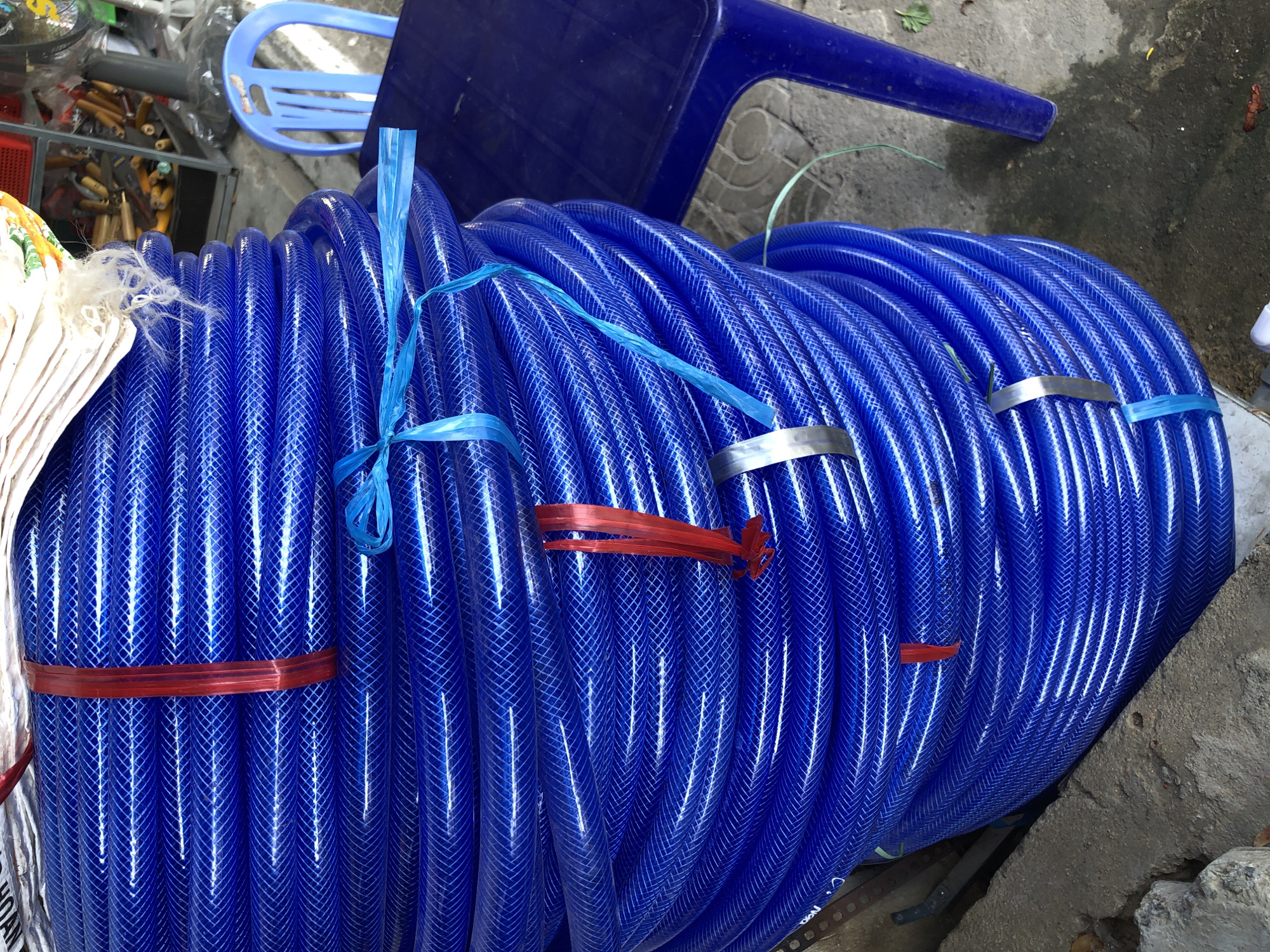 A large coil of blue, ribbed plastic hosepipe, banded with red, blue, and silver ties, is stacked vertically on a concrete surface. The hosepipe appears to be commercially prepared for sale. To the right of the hosepipe is a blue plastic chair and a blue plastic table. Behind the hosepipe, partially visible, is a metal shelving unit containing various items, including what appear to be tools with wooden handles. The scene is outdoors, with a concrete pavement and some rough concrete blocks in the foreground. The image seems to be taken at street level, possibly a marketplace or a shop selling outdoor equipment in Đà Nẵng, Vietnam.