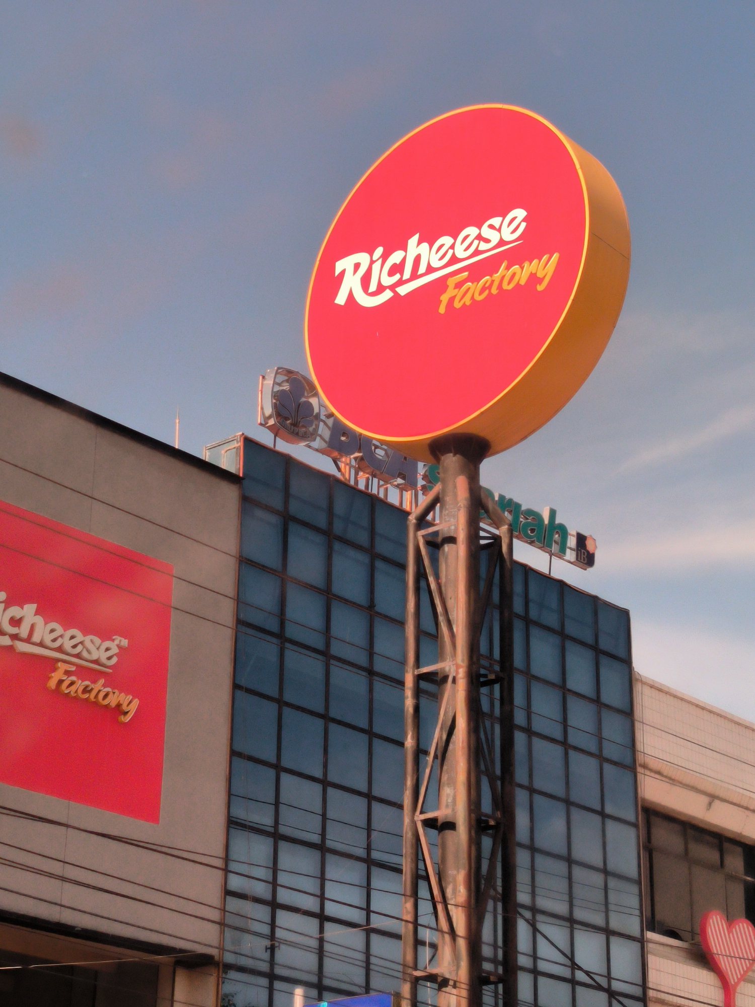 The image shows the Richeese Factory building in Lhokseumawe, Indonesia. The building has a prominent red and yellow circular sign on a metal support structure. The building itself has a large glass facade with a visible grid pattern, and an adjacent section with a lighter-colored, textured surface. A smaller Richeese Factory sign is affixed to the building's facade. The sky is visible in the background.