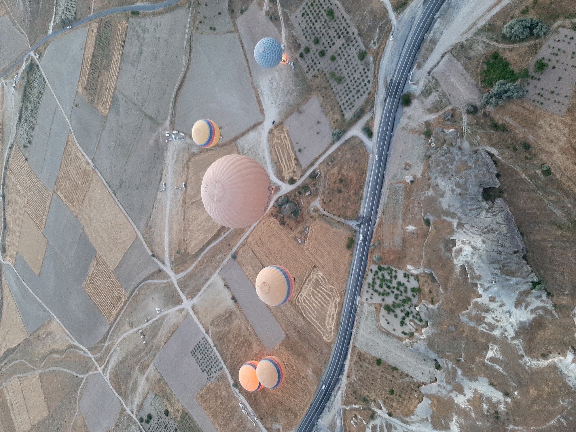 The aerial view captures a landscape that is characteristic of Cappadocia, near Avanos, Turkey. The terrain is a mosaic of agricultural fields, some fallow and others showing patterns of cultivation. A winding paved road runs through the scene, with a few white vehicles visible on it. Scattered across the landscape are several hot air balloons, appearing to be in the process of a flight. One large, pale orange balloon dominates the center, while other smaller balloons, some yellow with red and blue stripes and one light blue with a checkered pattern, are also visible. The ground shows signs of erosion and geological formations, with rocky outcrops and valleys. The time of day appears to be early morning or late afternoon, given the soft lighting.