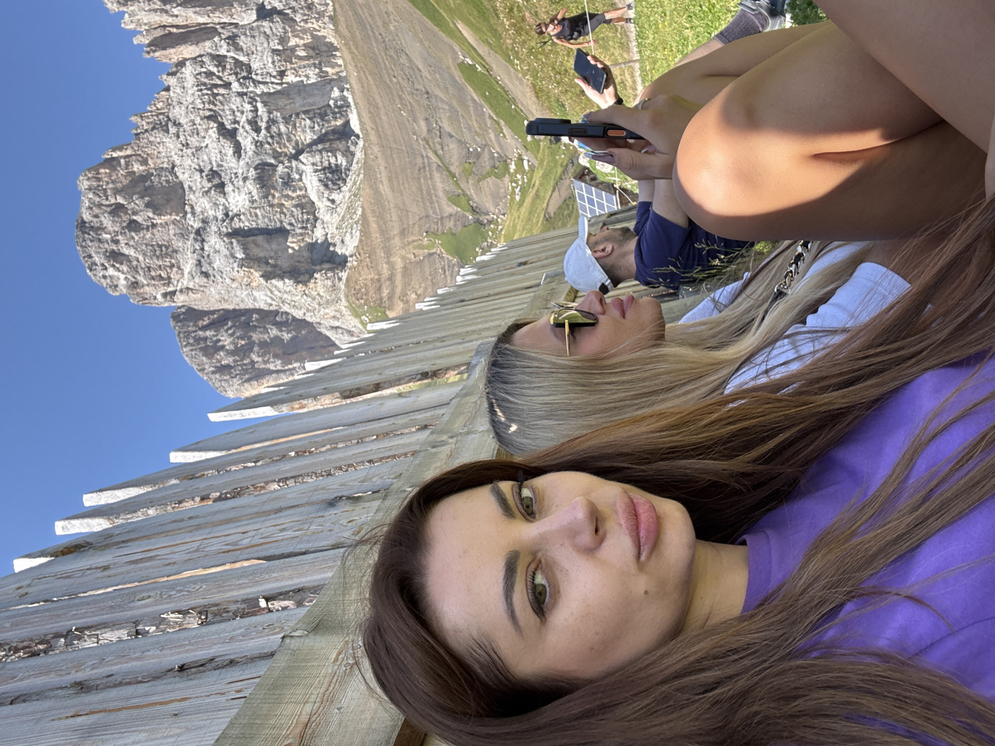 In an outdoor scene in Campitello di Fassa, Italy, three people are seated on a grassy slope, with a wooden fence in the foreground and a rocky mountain range in the background. A woman with long brown hair, wearing a purple t-shirt, is in the front, looking towards the camera. To her right, another woman with blonde hair, wearing sunglasses and a white t-shirt, is looking away from the camera. A person wearing a white cap and a dark blue t-shirt is seated behind them, holding a phone. In the distance, another person is walking. The environment is sunny with a clear blue sky.