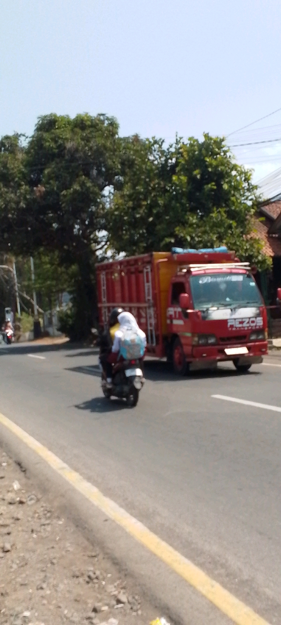In a sunlit scene on a paved road in Cilaku, Indonesia, a red truck with a yellow cargo box is stationary. The truck has "REZOS TRANSPORT" written on its front. To the left of the truck, a motorcycle travels away from the viewer. The motorcycle carries two passengers; the one in the rear wears a white headscarf and a backpack. The environment includes a tree line behind the truck, with lush green foliage. The road has a yellow line marking its edge on the left. Overhead, utility wires are visible against a pale blue sky.