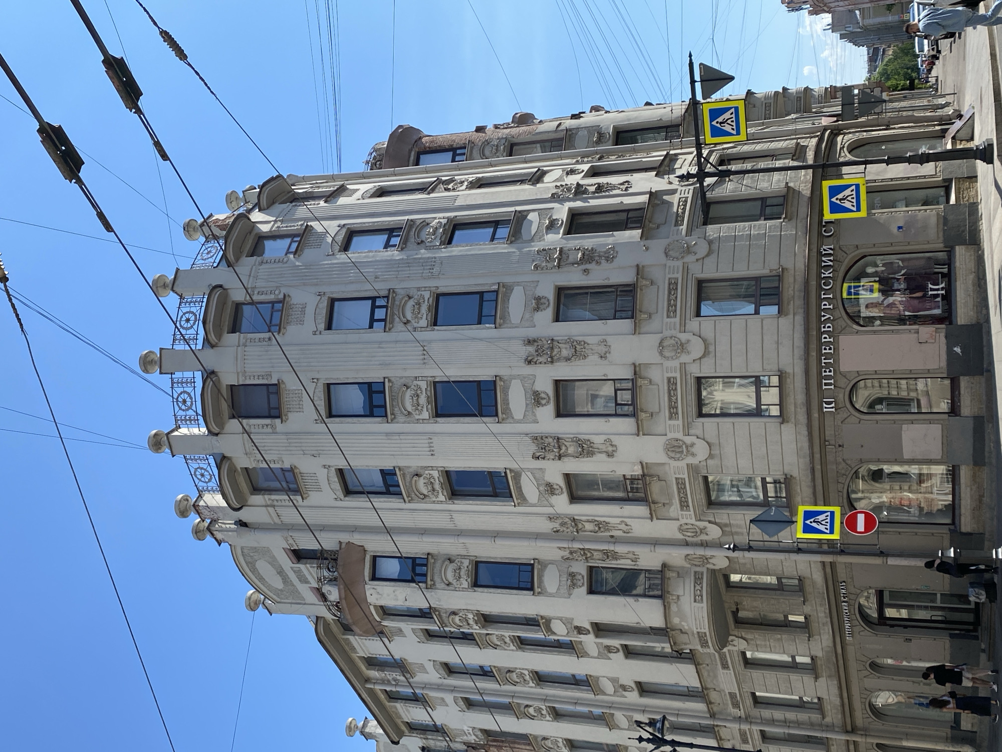 The image captures a vibrant daytime street scene in Saint Petersburg, Russia, focusing on an ornate, multi-story building under a clear blue sky. The structure, characteristic of Art Nouveau or Modernist architecture, features a light-colored facade extensively adorned with intricate stucco decorations, including elaborate reliefs, pilasters, and stylized garlands. Distinctive circular motifs are prominent on the small balconies and balustrades of the upper floors. The windows vary in shape and size, many with dark frames, reflecting the bright sky.
Numerous dark overhead wires, likely for public transport like trams or trolleybuses, crisscross the sunny blue sky, forming a dynamic element against the urban backdrop. At street level, the building houses commercial establishments; one large display window showcases mannequins, suggesting a clothing store. The building's facade bears Cyrillic text, reading "ПЕТЕРБУРГСКИЙ СТ" (Petersburg St.), likely part of a shop's name or a street reference.
On the paved sidewalk, a black pole holds two yellow square signs indicating a pedestrian crossing and a red circular 'No Entry' sign. Several casually dressed pedestrians are visible walking or standing on the sidewalk. The bright sunlight and cloudless sky indicate a clear, pleasant day. The perspective, looking up, emphasizes the building's height and its rich architectural detailing, offering a glimpse into daily life in Saint Petersburg.