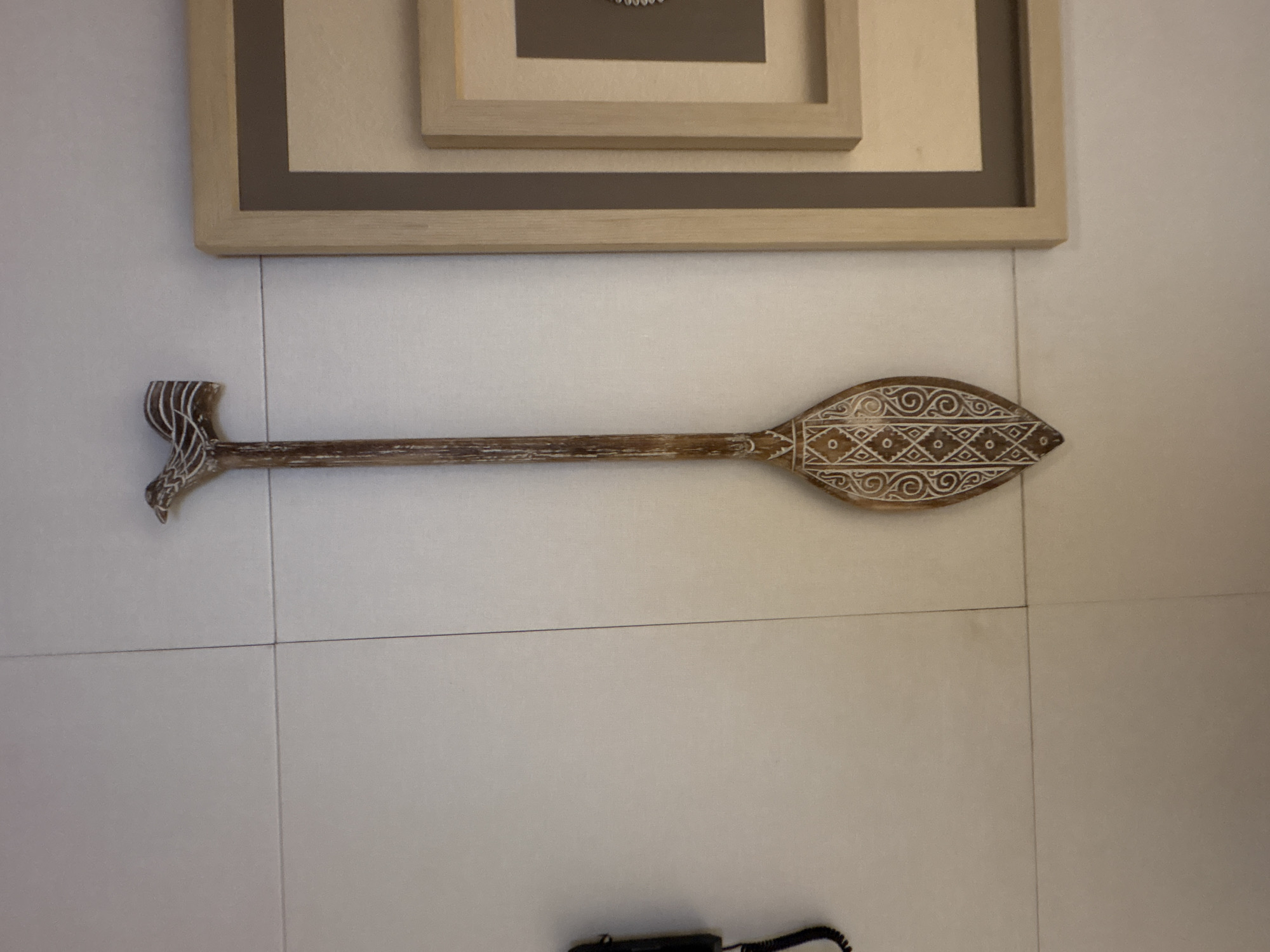 A wooden, carved decorative paddle is mounted on a tiled wall, likely in an interior space in Vagator, India. The paddle is elongated with a carved bird-like figure at the top and intricate geometric patterns along the wide, flattened end. To the right of the paddle, a framed artwork is also displayed on the wall. A black telephone receiver and cord are visible on the left side of the image. The wall appears to be made of large, light-colored rectangular tiles.