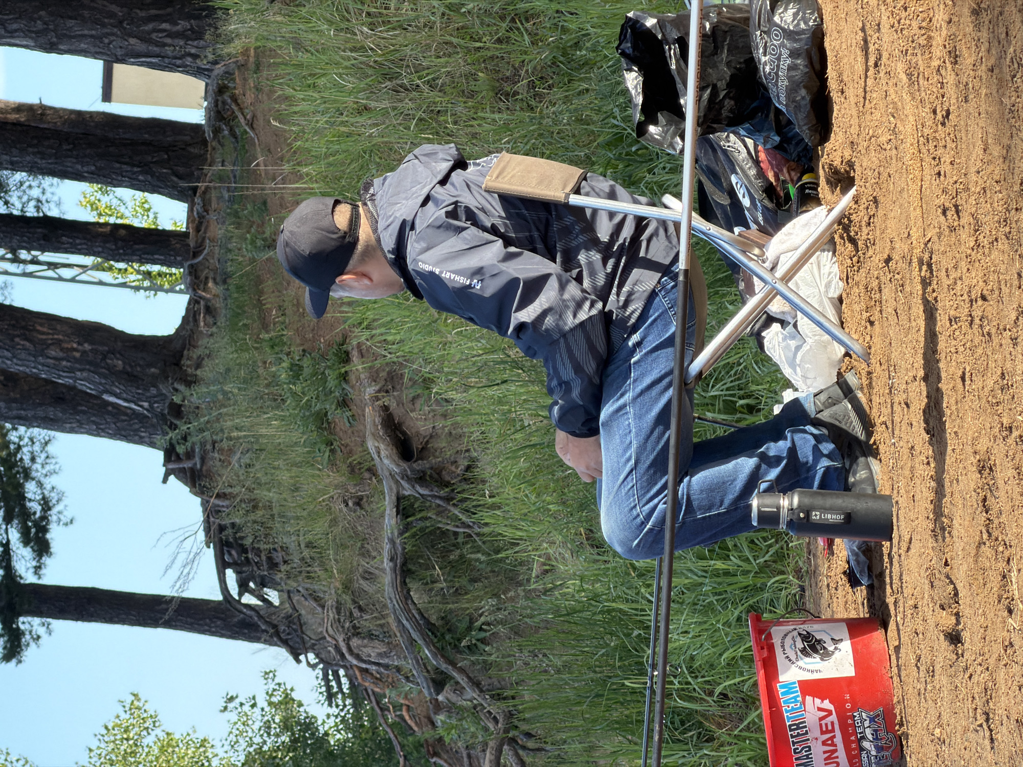 An outdoor scene depicts a person engaged in fishing on a bright, clear day, likely in Chaikovsky, Russia. The individual, a man, is seated on a portable folding chair on a patch of dry earth adjacent to green grass. He wears a dark baseball cap, a dark blue and grey jacket with a patterned design, and blue jeans. A light brown pouch is visible on the back of his jacket.
Two fishing rods extend horizontally across the foreground, indicating the primary activity. Beside the man's feet, a black thermos or water bottle with the brand "LIBHOF" is placed. A prominent red plastic bucket, adorned with several stickers, sits on the ground. These stickers display text such as "MASTERTEAM," "UNAEV," "WORLD CHAMPION," "RUSSIAN TEAM," and "ЧАНКОВСКИЙ РЫБОЛОВ." (Chaikovsky Fisherman), along with a stylized fish logo. Several black plastic bags, one partially showing "купки!" (meaning 'purchases' in Russian), are stowed near the chair. The man's jacket also features "F FISHART STUDIO" text.
The background reveals a natural landscape with a raised embankment covered in grass and large trees, possibly pines, whose roots are visible along the slope. The sky is bright blue, suggesting pleasant weather. The scene conveys a sense of calm and focused activity, characteristic of fishing.