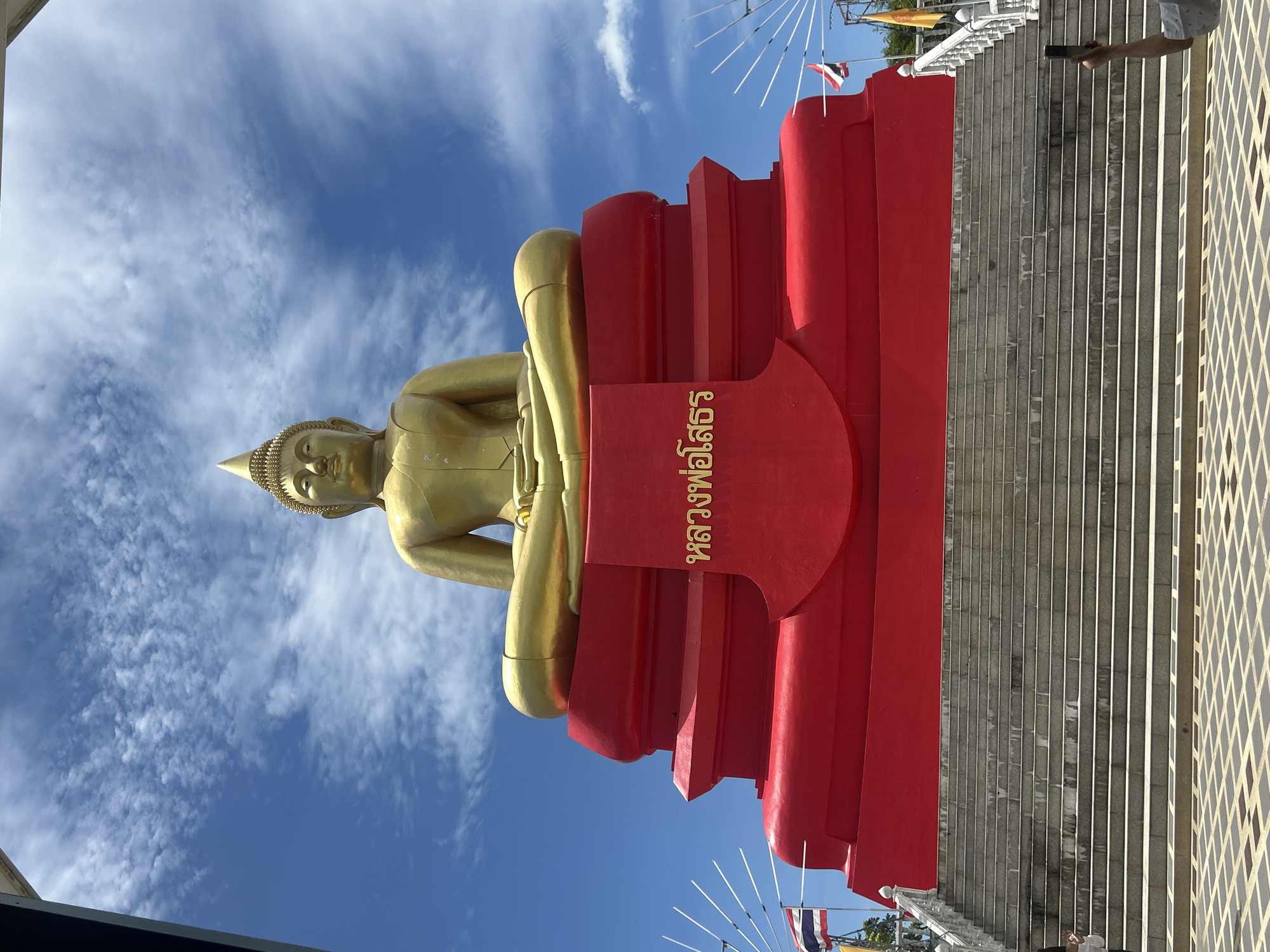 A large, golden Buddha statue is seated on a tiered, bright red platform. The statue faces forward, with its legs crossed in meditation. The platform has inscriptions in gold lettering, which appear to be in Thai script. Below the platform, a wide set of stone stairs leads down to a patterned paved area. The sky above is blue with scattered white clouds. Thai flags are visible on poles to the sides of the statue. A person's arm is visible on the right, holding a smartphone to take a picture. The image appears to be taken from a low angle, emphasizing the scale of the Buddha statue and its platform. The overall scene suggests a religious or tourist site, possibly a temple or shrine. This location is identified as Wat Phra Non Chantha Ram, located in Thai Ko Subdistrict, Mueang Phatthalung District, Phatthalung Province, Thailand.