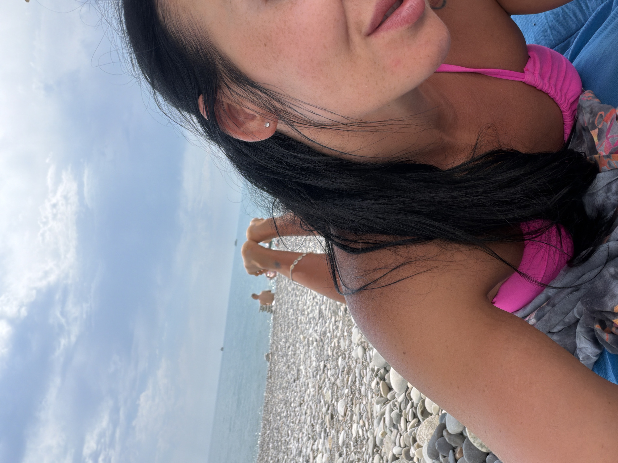 A young woman with long black hair lies on a pebbled beach in Sochi, Russia, her body partially visible from a low-angle perspective. She wears a bright pink bikini top. Her face is mostly out of frame, with only her cheek, ear, and mouth visible. She has freckles on her skin and a small stud earring in her left ear. Her arms are bent, and her hands are clasped together, resting on the pebbles. In the background, the calm sea stretches out to the horizon under a cloudy sky. A few other people are visible in the water and on the beach in the distance. The overall scene depicts a relaxed day at the beach.