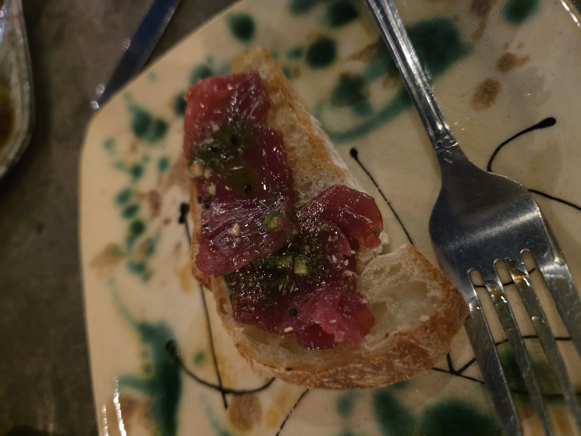 A close-up shot of a plate with a slice of bread topped with raw fish and green herbs. The plate has an abstract pattern of green, brown, and cream colors with black lines. A fork rests on the plate next to the bread slice. A knife and small dish are partially visible in the background. The dish might be located in Ensenada, Mexico.