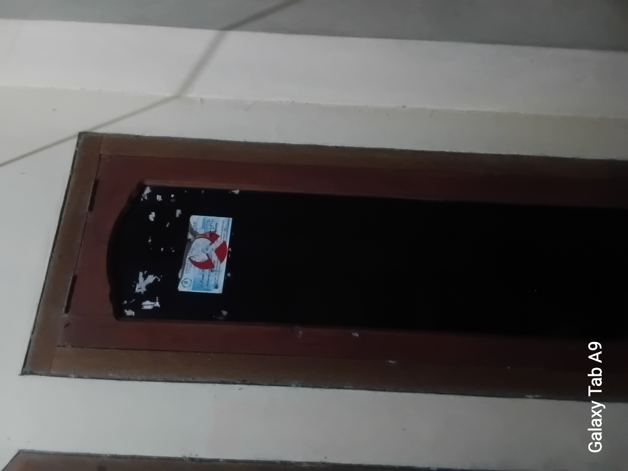 The image shows a closed wooden door with a dark opening. Two small stickers are affixed to the upper portion of the door, on the left side of the opening. One sticker is mostly obscured by glare and damage, but it appears to be a blue and white circular design with some text. The other sticker below it is more visible and shows an Arabic inscription with a red and white circular emblem. The door is set within a wall that appears to be painted a light cream color. The overall lighting suggests the photograph was taken in low-light conditions, possibly indoors at night. The bottom left of the image has a watermark that reads "Galaxy Tab A9". Without further context or identifying features, the specific location within Trenggalek, Indonesia cannot be determined.