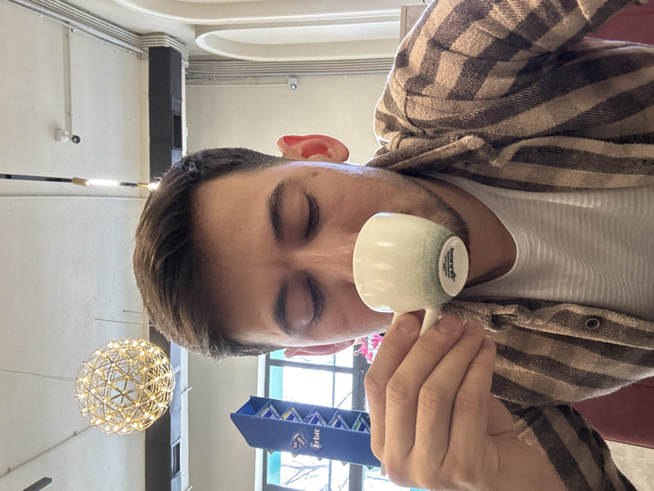 A person with closed eyes is holding a small, white and green cup close to their face. They're wearing a white shirt and a brown plaid jacket. A prosthetic ear is visible near their hairline. In the background, there's a blue display rack with the "Orbit" logo, a decorative light fixture resembling a globe, and a window. The photo appears to be taken in a public space, possibly a store or cafe.