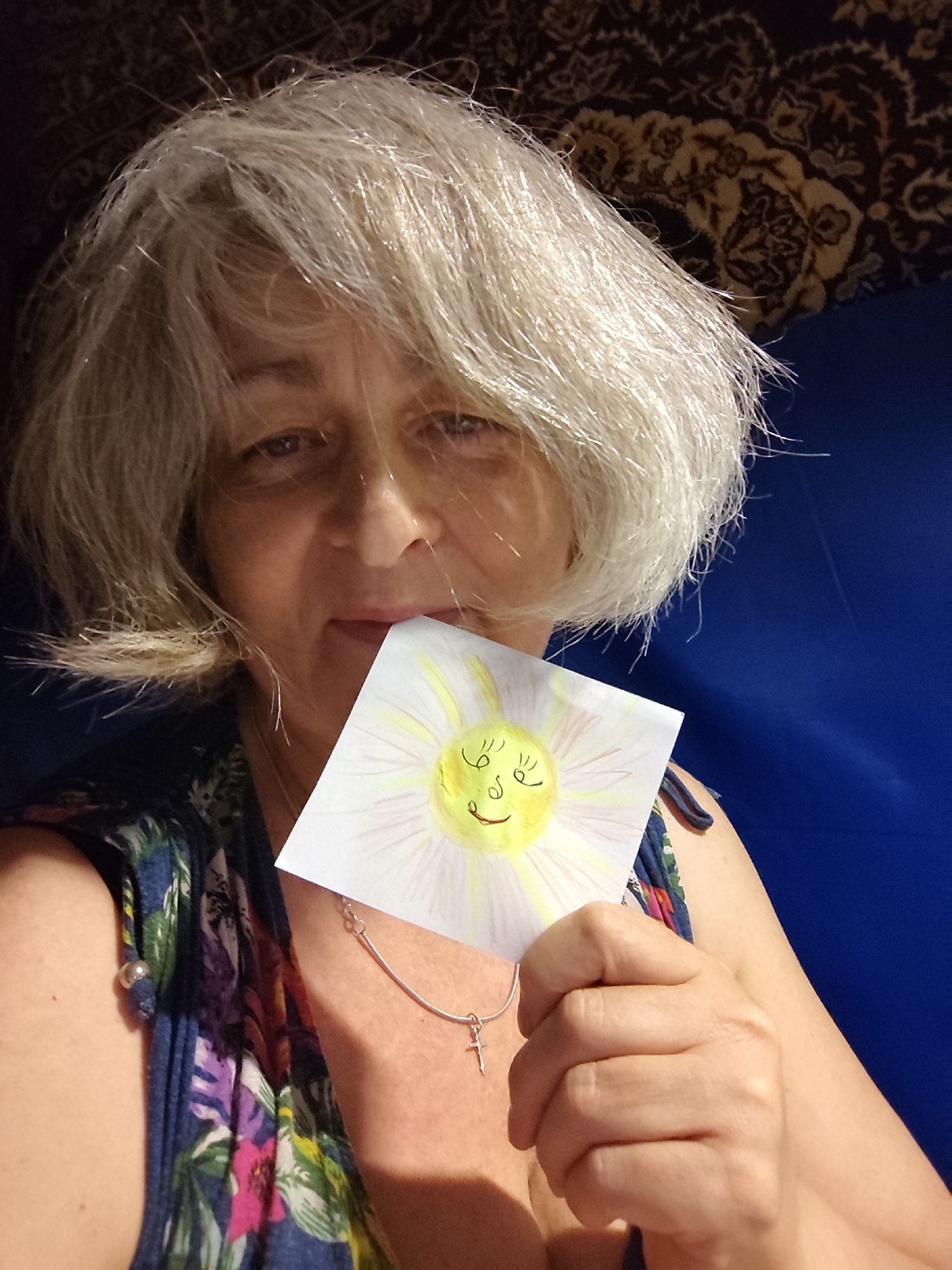 A medium shot captures a woman with short, light-colored hair holding a drawing of a smiling sun in front of her face. She is wearing a colorful floral print top and a silver necklace with a small cross pendant. The background consists of a dark, patterned fabric and a blue surface. The scene appears to be indoors, with artificial lighting. There is no indication of the specific location in Krasnodar, Russia.