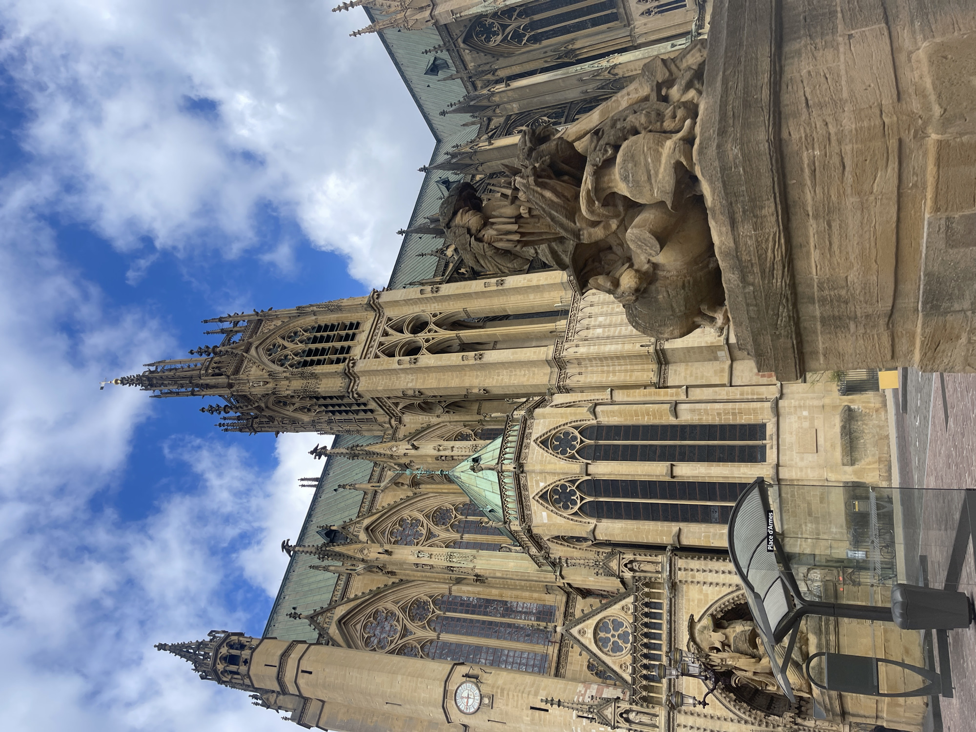 The image depicts the Metz Cathedral, formally the Cathedral of Saint Stephen of Metz, a prominent landmark in the city of Metz, France. The photograph is taken from a low angle, accentuating the cathedral's Gothic architecture, including its intricate spires, ornate stone carvings, and large stained glass windows. The structure is crafted from light-colored stone, contrasting with the partially visible blue sky dotted with white clouds in the background. A modern bus stop shelter, made of glass and metal, is situated in the foreground, with "Place d'Armes" visible on the glass. A stone statue with a weathered appearance is positioned prominently on the cathedral’s exterior. The overall impression is one of historical grandeur juxtaposed with modern urban elements.