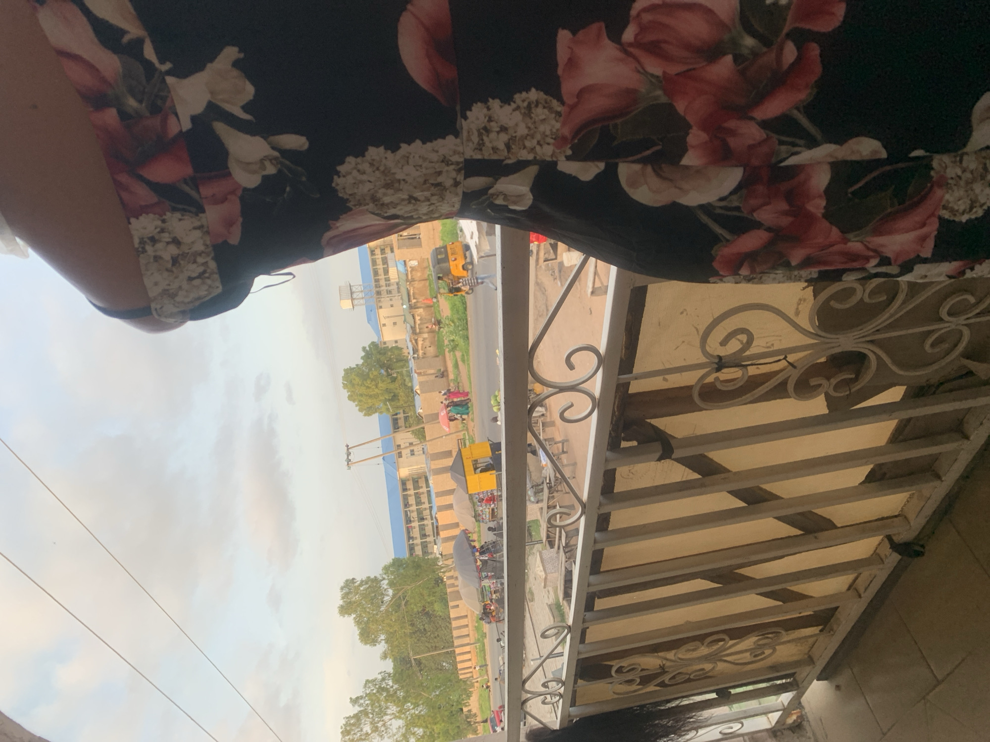 The image depicts a view from a balcony overlooking a street scene in Jalingo, Nigeria, likely during the late afternoon given the lighting. In the foreground, a portion of a person wearing a dark floral dress is visible, obscuring the right side of the frame. The balcony railing, made of metal with decorative scrollwork, runs horizontally across the lower part of the image.
Beyond the railing, a street is lined with market stalls and various vehicles. A bright yellow auto-rickshaw, commonly known as a "Keke Napep," is prominent in the middle ground, along with several other vehicles, including a red car. Pedestrians are scattered throughout the scene, some interacting with vendors at the market stalls, which are covered by umbrellas. In the background, buildings, including a school with a blue roof and a water tower, are visible. Lush green trees are also interspersed throughout the urban landscape. The sky is overcast with some patches of blue, indicating a mixture of clouds and sunshine.