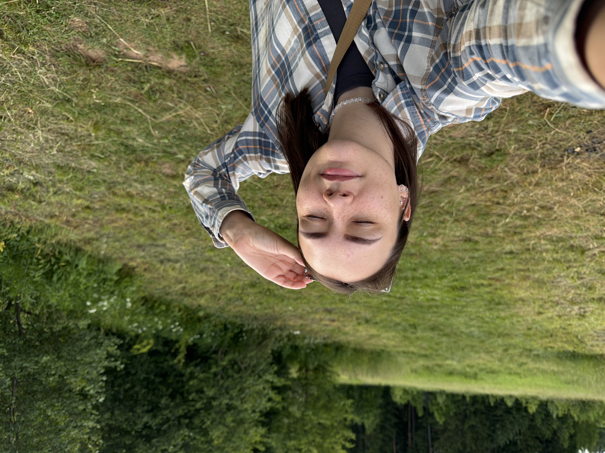 A young woman with brown hair and freckles is taking a selfie in a grassy, wooded area. She is wearing a plaid shirt and has her eyes closed, with her hand on her forehead. The background is a blurred landscape of trees and grass. The location is Garevaya, Russia.