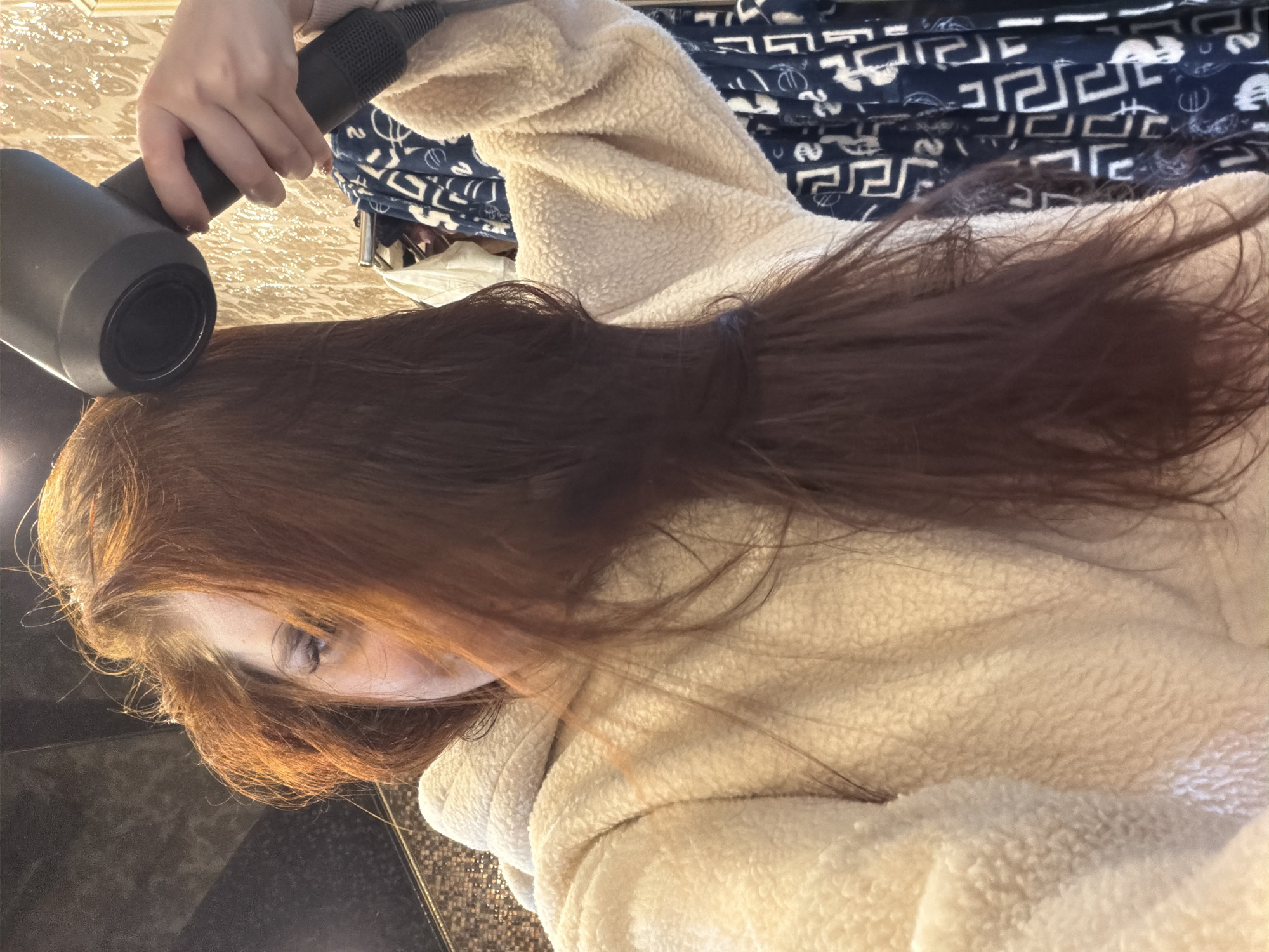A person is drying their long, dark brown hair with a black hairdryer. The person is wearing a fuzzy, cream-colored robe. The background includes a patterned blue and white fabric, possibly a blanket or clothing, and a textured surface that appears to be a countertop. The lighting suggests it could be daytime, but the exact time of day and weather are not discernible. The scene appears to be indoors, likely in a bathroom or bedroom. The person's face is partially visible, but their expression is not clear. There is no visible text in the image.