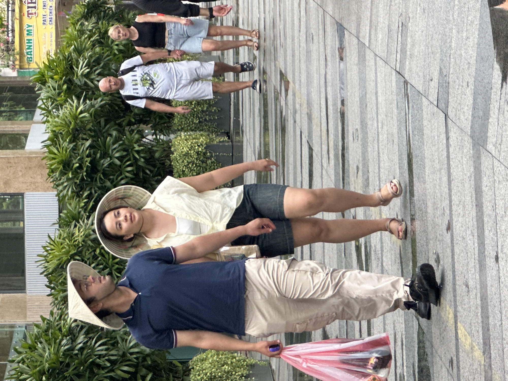 The image depicts a street scene in Da Nang, Vietnam, likely in the vicinity of a commercial area given the signage visible in the background. In the foreground, two couples are walking on a paved sidewalk. The couple on the left consists of a man wearing a blue polo shirt, khaki cargo pants, and a conical hat, carrying a pink plastic bag. Beside him, a woman also wears a conical hat, a light-colored blouse, and denim shorts. The couple on the right comprises a bald man in a white "Jordan" t-shirt and grey shorts, and a woman in a black tank top and denim shorts. They are holding hands as they walk. The environment is urban, with lush green foliage lining the street and reflections on the wet pavement suggesting recent rain. The overall impression is of a casual stroll through a city.