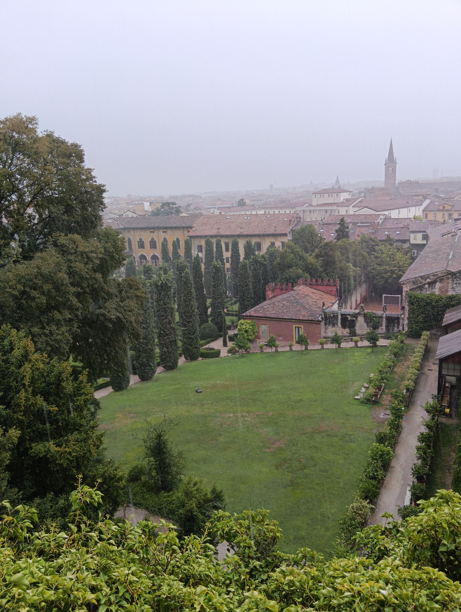 The image depicts a view of the Giardino Giusti in Verona, Italy, on a misty or rainy day. In the foreground, lush green foliage dominates, with the leaves of a large tree on the left side of the frame appearing wet. Beyond the foliage, a well-maintained rectangular lawn occupies the center of the image.
Behind the lawn, a path lined with manicured bushes and trees leads to a rustic, reddish-brown building with a tiled roof. This building appears to be part of the garden complex. Further back, more trees, including a row of tall, slender cypress trees, are visible.
In the middle ground, a large, palatial building with arched windows and a yellow facade stands behind the cypress trees. The background reveals a dense urban landscape of Veronese rooftops, characterized by terracotta tiles, under a hazy, overcast sky. A prominent clock tower, characteristic of Verona's skyline, rises above the buildings in the distance, partially obscured by the mist. The overall atmosphere is subdued, suggesting a cool, damp day. There are no people visible in the scene. The weather appears to be a light rain or mist, as evidenced by the droplets on the foliage and the softened light.