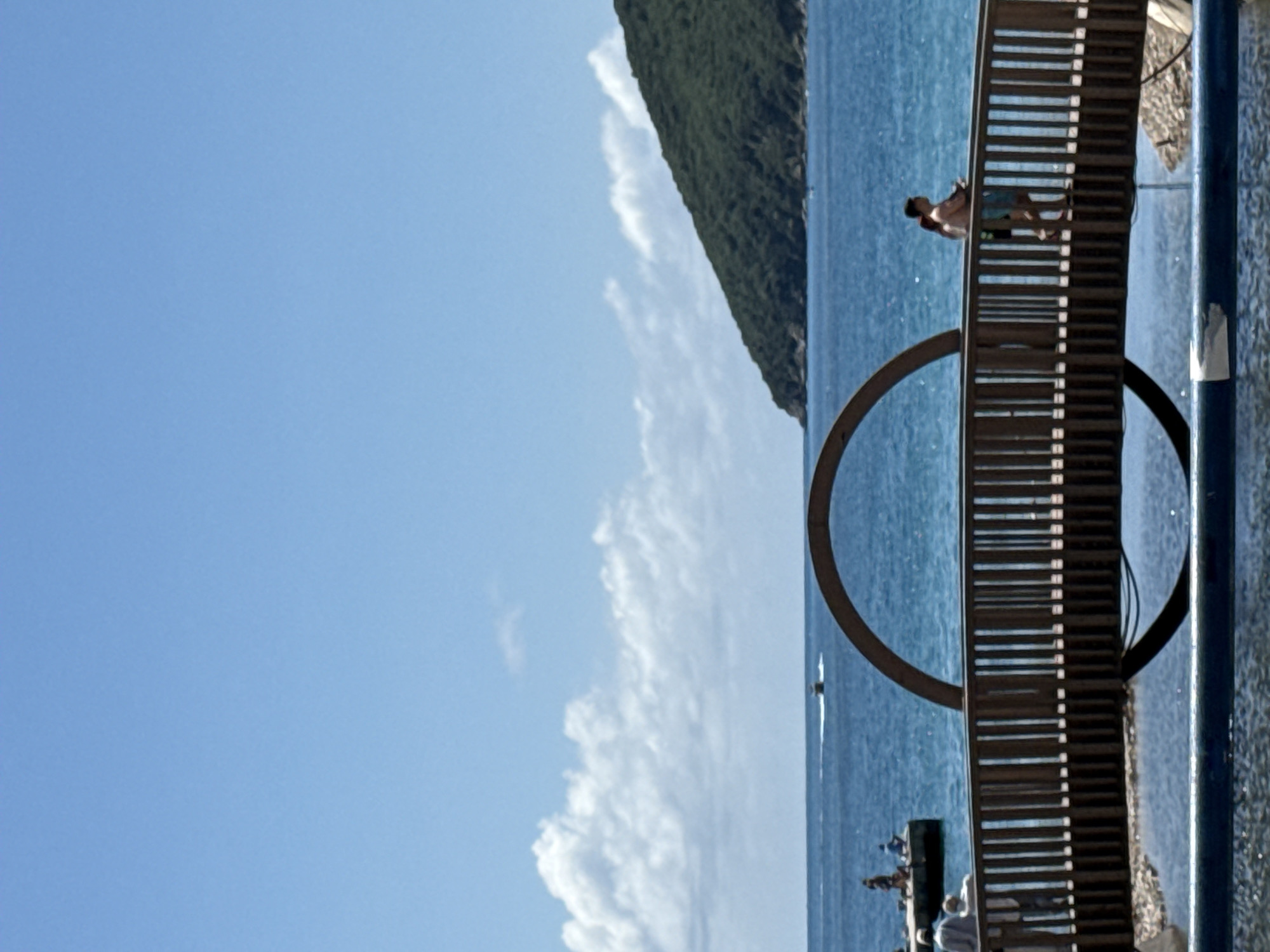 The image presents a vibrant, sunlit coastal scene in Budva, Montenegro. The composition is split between the expansive blue sky with scattered white clouds on the left and the deep blue sea on the right, separated by the distant silhouette of a green, tree-covered mountain range.
A prominent wooden walkway or pier extends into the sea, forming a key feature in the mid-ground. Towards the upper right of this structure, a man wearing swim shorts stands, seemingly enjoying the view or preparing for an activity in the water. Directly in front of the walkway, a striking large, dark circular sculpture or archway emerges from the sea.
Further in the distance on the water, a small boat or jet ski is visible, creating a faint white trail. To the lower left of the main walkway, a small group of people can be seen on a lower platform or dock, also enjoying the waterfront.
The bright sunlight, clear sky, and calm sea indicate a warm, pleasant day, likely suggesting summer. The scene evokes a sense of leisure and natural beauty, characteristic of a coastal resort area. There is no visible text in the image.