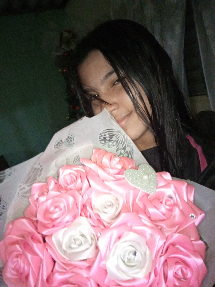 A young woman with long, dark hair smiles while holding a bouquet of pink and white satin ribbon roses. The roses are arranged in a close bunch, with a small glittery heart attached to one. A white paper with butterfly patterns partially covers the woman's face. The background includes a green wall on the left and a window with light-colored curtains on the right, and a small decorated Christmas tree is visible in the background.