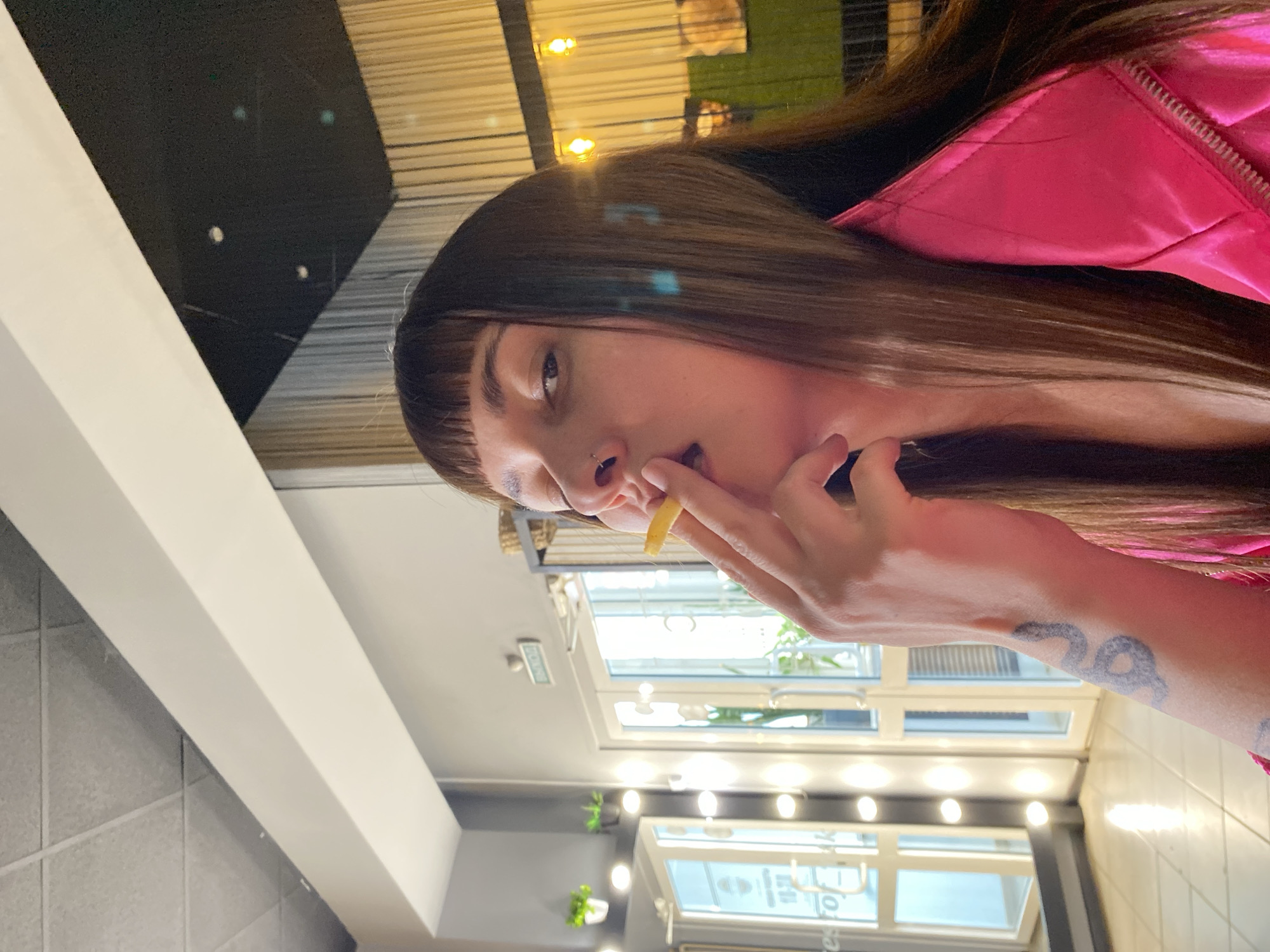 The image captures a person, likely a woman, with long, straight brown hair and bangs, looking directly at the camera. She has a small stud nose piercing. She is wearing a bright pink, possibly padded or quilted, jacket. She holds a single french fry to her mouth with her right hand, appearing to be in the process of eating. A distinctive tattoo, which appears to be a stylized design or numbers (possibly "20" or "29" or similar script), is visible on her right wrist.
The setting is indoors, likely a cafe or restaurant, characterized by light grey tiled walls and white architectural beams or trim. Large windows or glass panels form a significant part of the background, reflecting multiple bright, round indoor lights. Through the glass, some indistinct outdoor elements and small potted plants arranged on shelves are visible. Partially visible reflected text can also be seen on the glass. The predominant lighting is artificial, suggesting an indoor ambiance during evening or night, or a well-lit interior space. The photograph is taken at a tilted angle.