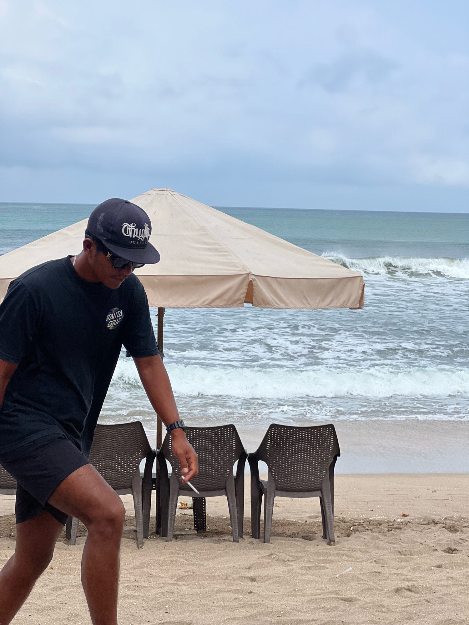 In Kuta, Indonesia, a man walks on a sandy beach near the ocean under an overcast sky. He is wearing a black t-shirt, black shorts, a black baseball cap with white lettering, and sunglasses. He holds a cigarette in his right hand. Behind him, a large beige beach umbrella shades two brown plastic chairs. The ocean has moderate waves, and the water is a greenish-blue color. The sand appears wet near the waterline, indicating recent tide activity.