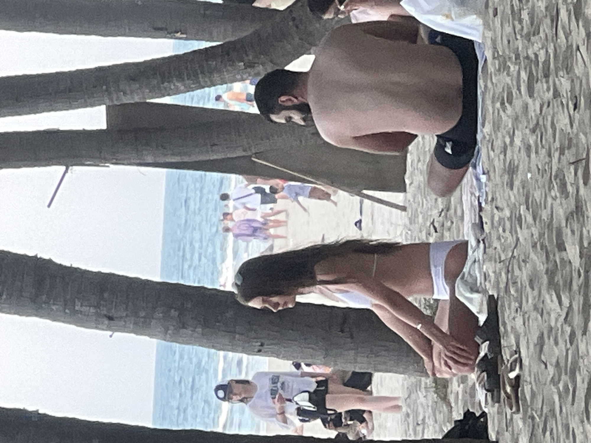 The media file displays a scene at a beach, likely Đà Nẵng, Vietnam, given the context. Several people are visible, most of whom are out of focus. In the foreground, two palm tree trunks are prominent, dividing the frame.
Beneath the palm trees, a woman is seen from the side, wearing a white bikini and with her dark hair flowing. She appears to be kneeling or bending down. To her right, a pair of flip-flops rests on the sand.
Further in the background, a man with dark hair is partially visible, seated and looking away from the camera. He is wearing swim trunks. Other individuals can be seen in the distance, near the water, some wearing swimwear. The ocean is visible in the background, with calm blue water under a bright sky, suggesting it is daytime with good weather. The sand is textured and appears light in color. There is no visible text in the image. The overall mood seems relaxed and casual, typical of a beach day.