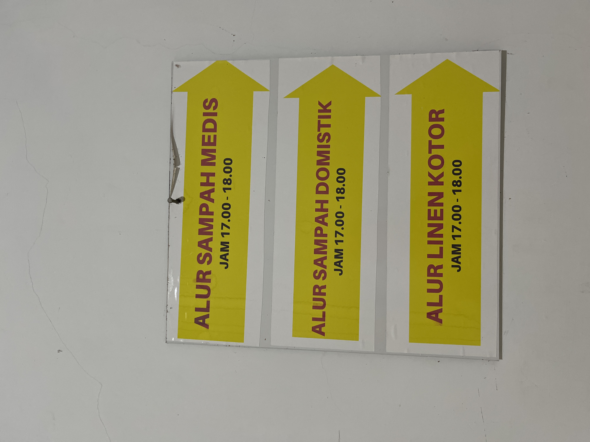The image shows a white wall with three signs stacked vertically. Each sign has a yellow arrow pointing to the right. The text on the signs is in Indonesian and indicates different types of waste disposal and their operating hours.
The top sign reads "ALUR SAMPAH MEDIS JAM 17.00 - 18.00," which translates to "MEDICAL WASTE FLOW 5:00 PM - 6:00 PM."
The middle sign reads "ALUR SAMPAH DOMISTIK JAM 17.00 - 18.00," which translates to "DOMESTIC WASTE FLOW 5:00 PM - 6:00 PM."
The bottom sign reads "ALUR LINEN KOTOR JAM 17.00 - 18.00," which translates to "DIRTY LINEN FLOW 5:00 PM - 6:00 PM."
The signs appear to be informational notices, likely posted in a facility such as a hospital, clinic, or hotel in the city of Lhokseumawe, Indonesia, given the language used. There are no people or actions depicted in the image. The environment is a plain white wall with some minor imperfections, possibly paint cracks or smudges. The signs are attached to the wall with small pins or staples at the top.