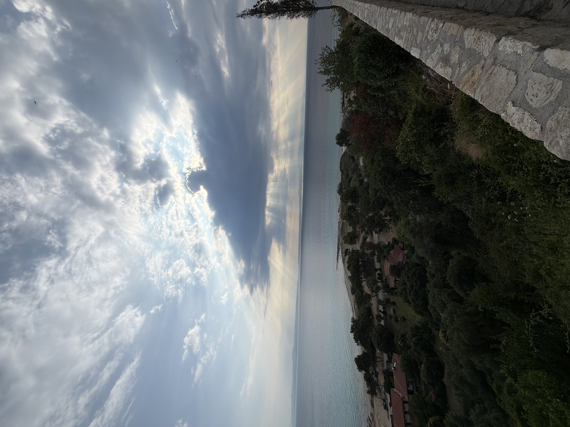 This is an outdoor, vertical, medium-shot, eye-level photograph of a coastal landscape at dusk. The scene depicts a body of water, possibly the Aegean Sea, stretching to the horizon under a dramatic sky filled with clouds. Sunbeams are breaking through the clouds, creating a luminous effect. In the foreground, there is a stone wall, and beyond it, a dense green hillside covered in trees and vegetation. Several houses with red tiled roofs are visible nestled amongst the trees. A beach with parked cars can be seen along the shoreline. The overall impression is one of a serene, natural setting with signs of human habitation. The location is likely the Municipal Unit of Kassandra in Greece, a region known for its coastal beauty.