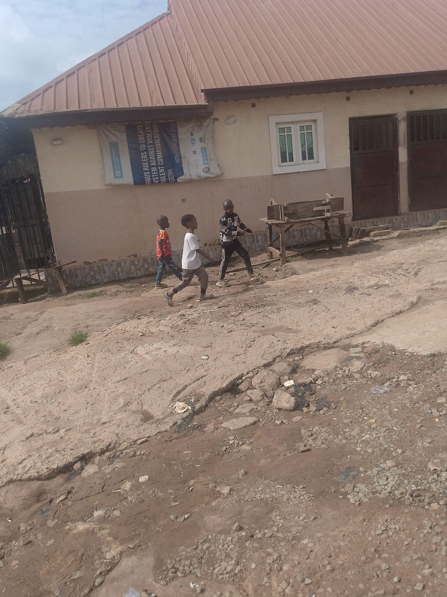 A group of three young boys walk along a dirt path in front of a building in Kurunduma, Nigeria. The building has a light-colored facade with a brown, sloped roof. There is a large banner hanging on the front of the building. A window with white panes and a brown, double-door entrance are visible on the right side of the building. In the foreground, the ground is uneven and appears to be a mix of dirt, rocks, and some debris. The sky is overcast.