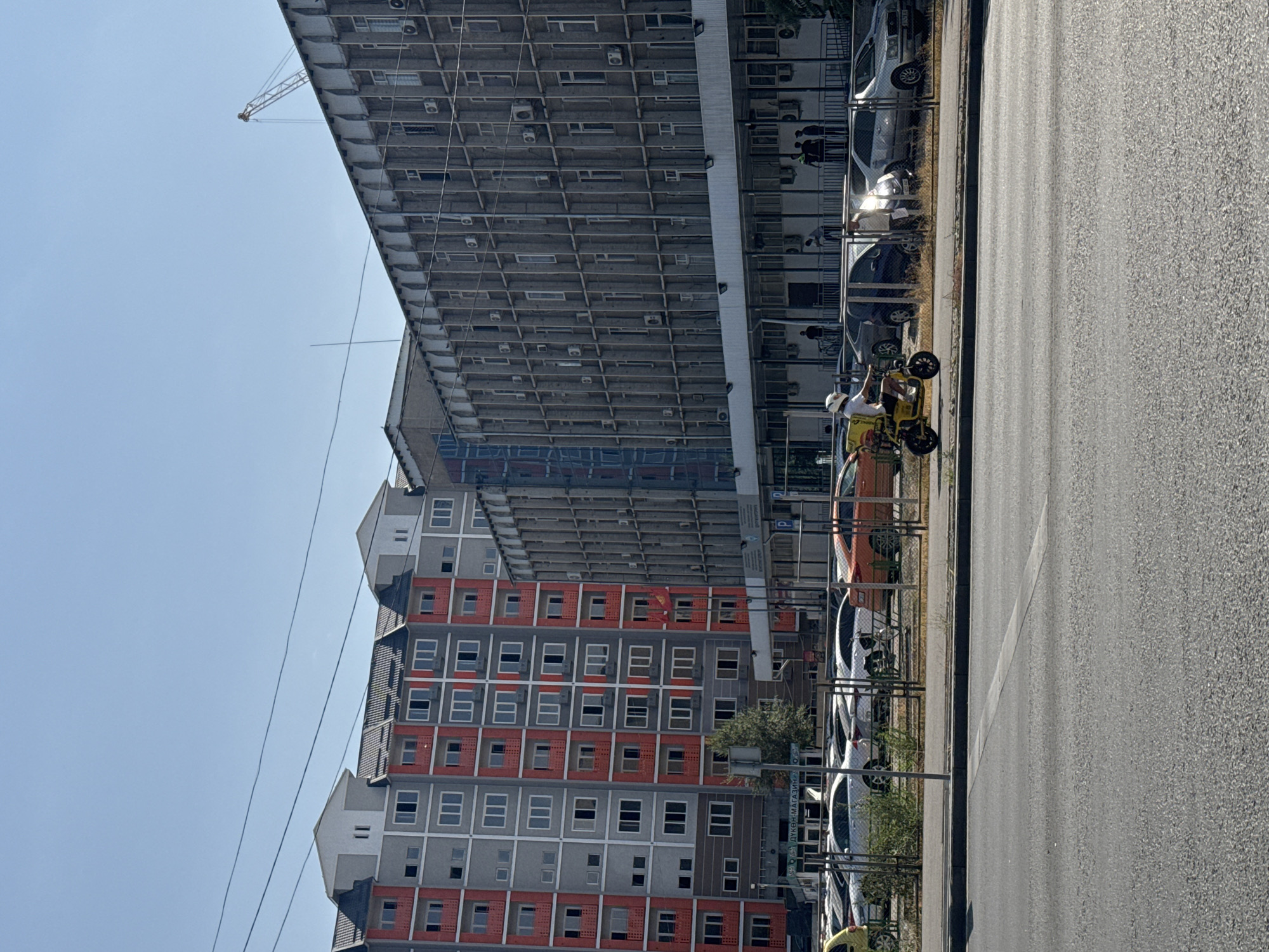 The image shows a street scene in Bishkek, Kyrgyzstan, on a clear, sunny day. In the background, a modern apartment building with grey and red facades stands next to a large, older concrete office building. A construction crane is visible above the office building. In the foreground, a person on a yellow electric scooter is riding on the street, heading towards the right of the frame. Several parked cars are visible behind a chain-link fence. The street appears to be a multi-lane road with clear pavement markings. The overall impression is of a bustling urban environment.