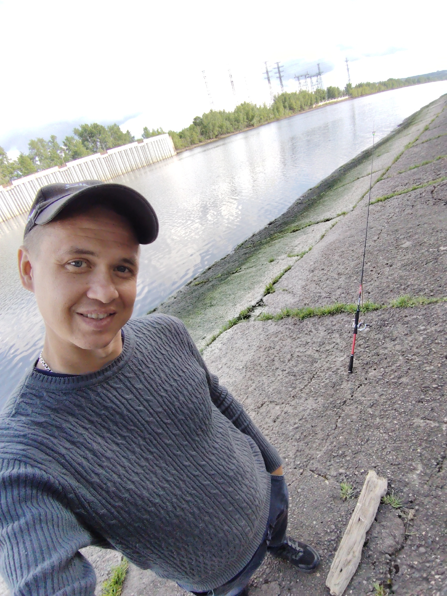 A man takes a selfie at a riverbank in Chaikovsky, Russia. He is wearing a gray sweater, a black baseball cap, and black boots. He is standing on a gray concrete bank next to a river. A fishing rod is propped against the bank behind him. In the background, there is a power plant and trees. A piece of driftwood lies on the ground.