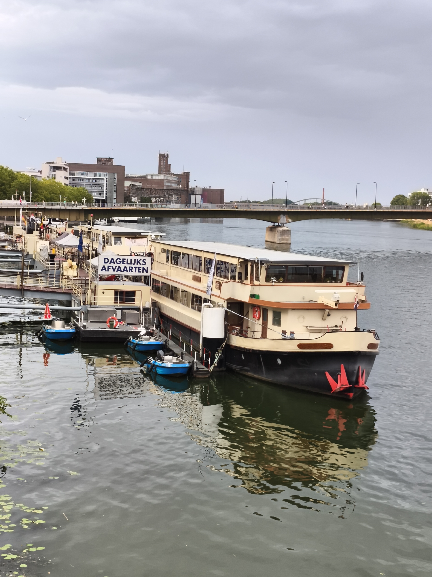 The image shows a quay in Maastricht, Netherlands, with several boats docked in a river under a cloudy sky. A large, light-colored passenger boat is docked prominently in the foreground, with the words "DAGELIJKS AFVAARTEN" (daily departures) displayed on a sign attached to it. Several smaller blue boats are also docked alongside the larger vessel. In the background, a wide bridge spans the river, with buildings and trees visible on the distant shore. The water is calm, with reflections of the boats and the sky visible on its surface. There are no people clearly visible in the image.