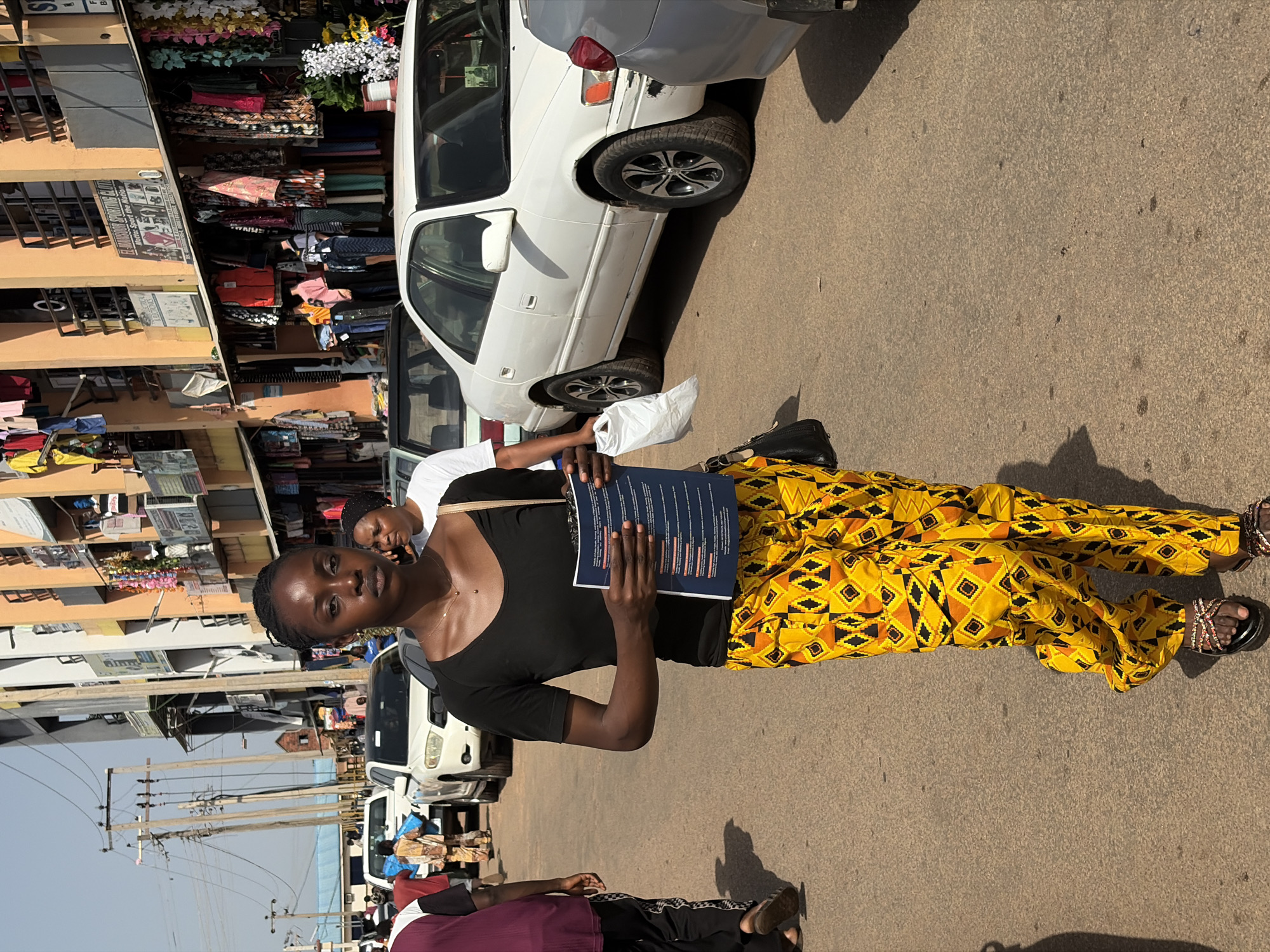 A young Black woman with braided hair walks towards the viewer on a dusty street in Jos, Nigeria. She wears a black, short-sleeved V-neck shirt and vibrant yellow pants with a geometric pattern. She holds a blue document in her right hand and a white plastic bag in her left. Her black shoulder bag is slung over her right shoulder.
The street is lined with buildings displaying various storefronts and goods for sale. Clothing racks with colorful garments hang outside the shops. Several white cars are parked or driving on the street. In the background, utility poles with a dense network of wires are visible. The overall scene depicts a busy market or commercial area.