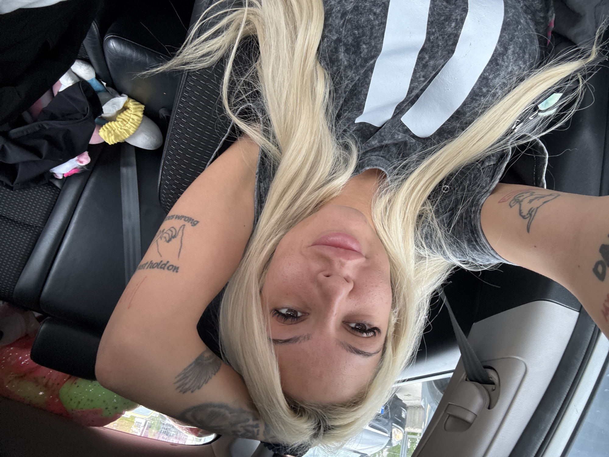A selfie captures a woman lying in what appears to be the front passenger seat of a car. She has long, straight blonde hair that falls around her face and shoulders. Her head is tilted slightly back, and she looks directly at the camera with a neutral, relaxed expression. She has defined eyebrows, dark eyelashes (possibly extensions), and her lips are subtly tinted pink.
She is wearing a dark grey t-shirt with a washed-out appearance and several prominent white vertical stripes or block letters on the front, possibly forming part of a logo or text. Numerous tattoos are visible on her arms. On her right forearm (viewer's left), there's a tattoo of two interlocking pinky fingers and text that reads "wrong" and "just hold on." Further down on the same arm, another tattoo depicts a pair of wings. On her left forearm (viewer's right), a tattoo resembling a dog's head (possibly a pitbull) is visible.
The interior of the car is dark, with black leather seats and a visible seatbelt mechanism on the right side of the frame. In the background, behind the woman's head, there's a jumble of items on the car seat, including dark clothing, a yellow scrunchie, and a grey, plush item. Partially visible in the lower-left corner is a plastic bag containing colorful, possibly round items. Outside the car, through the windows, a blurred street scene with other vehicles suggests it is daytime. The location context provided is Sofia, Bulgaria.