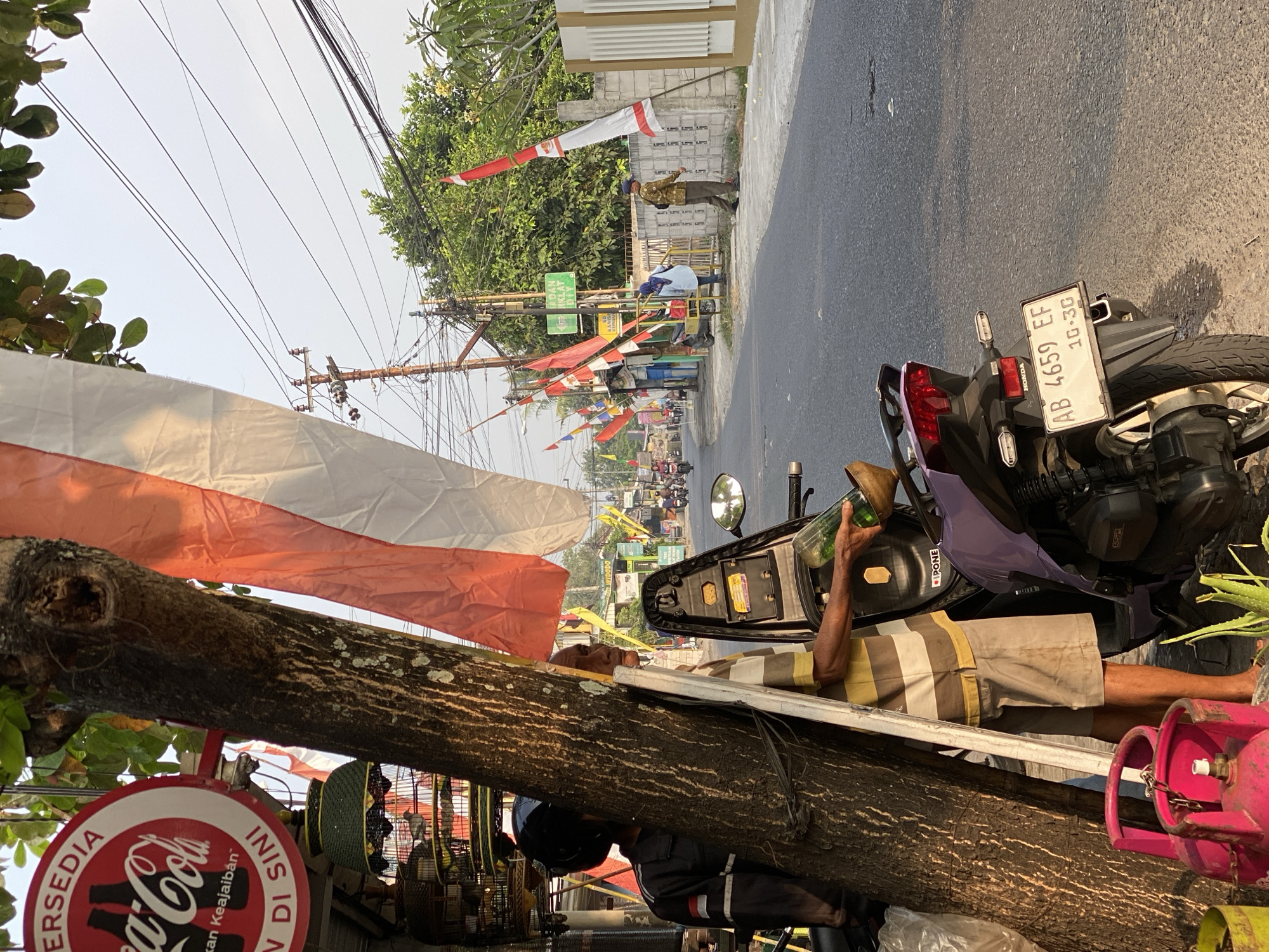 In Kasihan, Indonesia, along a street lined with trees and power lines, a man in a striped shirt and khaki shorts stands near a motorcycle, holding a green glass bottle and a wooden bowl. The scene is decorated with red and white Indonesian flags, suggesting a patriotic or celebratory occasion. A Coca-Cola sign is visible on a building in the background, along with birdcages and other vendor stalls. The street is paved, and the sky is clear. The time appears to be late afternoon, with the sun casting long shadows.