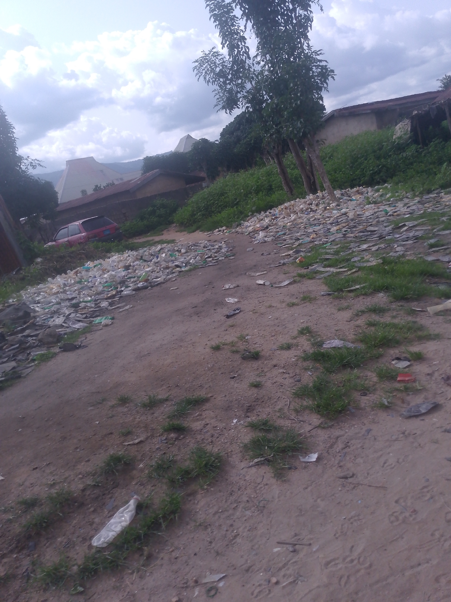 In Serti, Nigeria, the image depicts an outdoor scene characterized by a significant amount of scattered debris, primarily plastic bottles and other waste materials, strewn across a dirt path. The path is uneven and appears to be a natural or unpaved area, with sparse patches of grass growing between the waste. In the background, several structures are visible, including houses with corrugated metal roofs and what appear to be larger, possibly institutional buildings with unique geometric roof designs. A red car is parked on the left side of the path, partially obscured by vegetation and debris. Trees with full foliage are present, with some growing more densely near the background structures and others standing as solitary figures along the path. The sky is overcast with clouds, suggesting a diffused natural light. The overall impression is one of neglect and environmental disorder in a semi-urban or rural Nigerian setting.