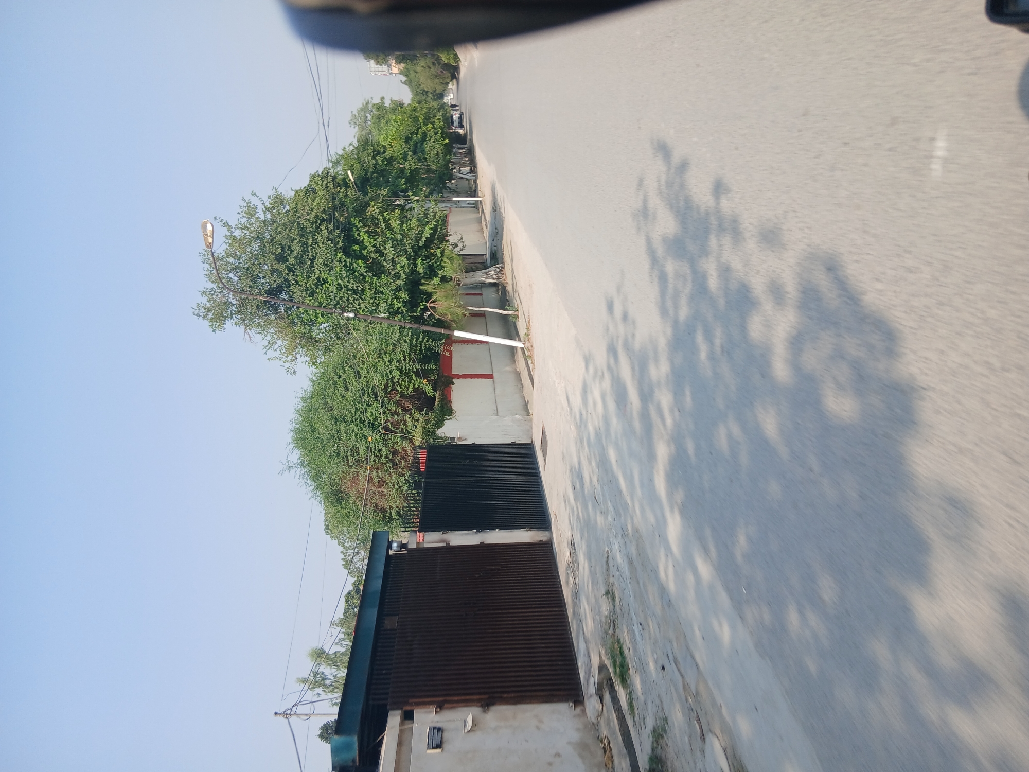 This image shows a street scene in Peshawar City Tehsil, Pakistan. On the left, there is a concrete wall with two large gates. The first gate is made of dark corrugated metal, and the second is a black metal gate. Behind the wall and gates are trees and a building with a green roof. A street light is visible above the trees. The street in front of the wall is paved and shows the dappled shadows of trees. The street continues into the distance, lined with more trees and some buildings. A car is parked on the side of the road further down. The sky is clear and blue. The overall impression is a residential or commercial area.